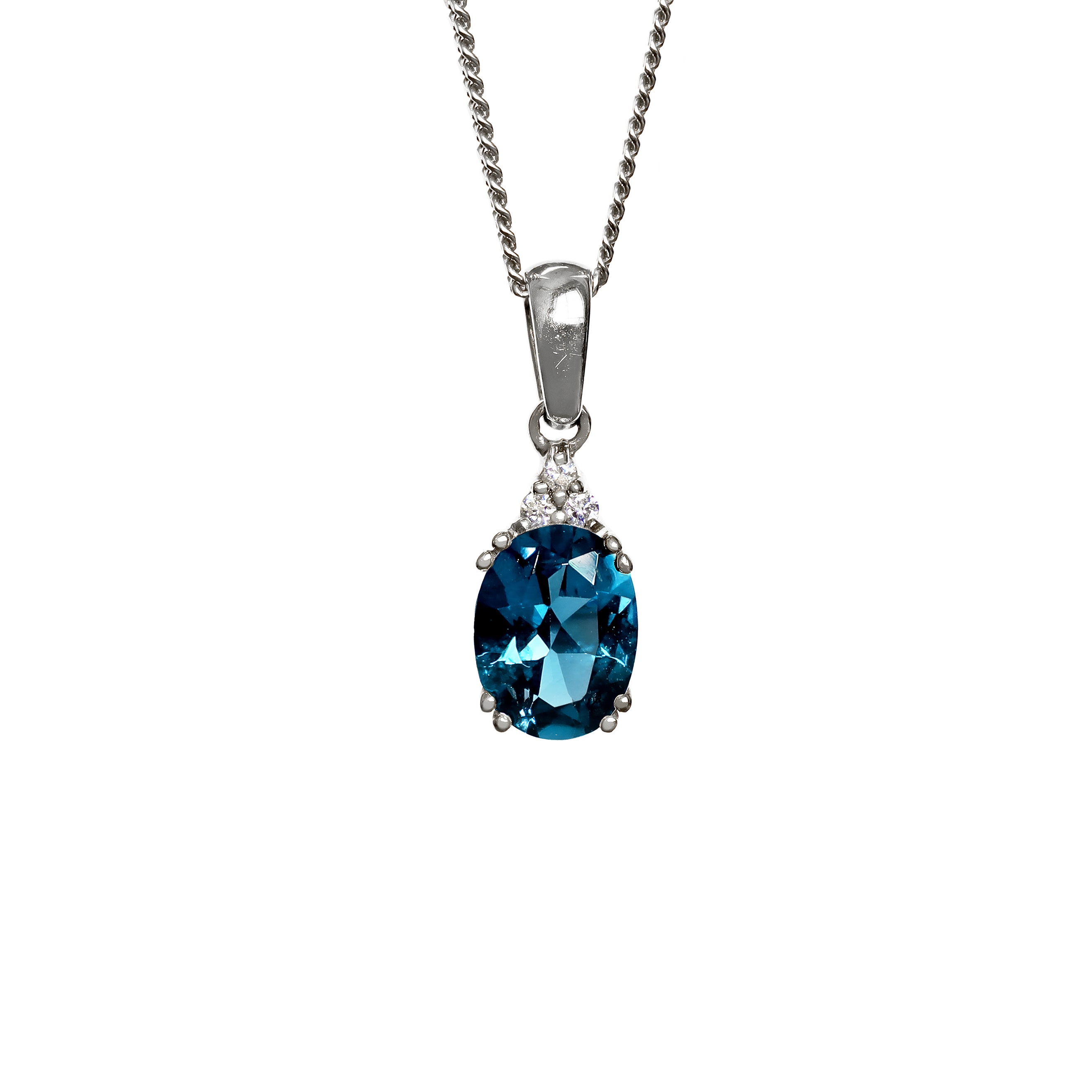 A product photo of a white gold london blue topaz pendant sitting against a white background. The deep blue oval-cut stone is held in place by 4 pairs of claws at its top and bottom, with a trio of white diamonds connecting the stone to the rest of the pendant. It is suspended by a simple white gold chain.