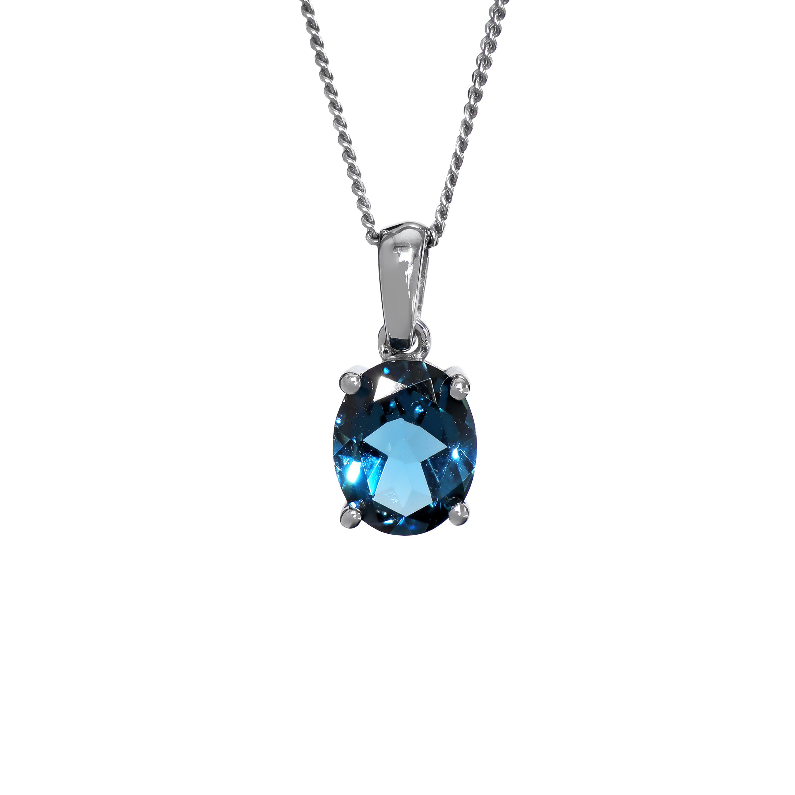 A product photo of a white gold london blue topaz pendant sitting against a white background. The impressively large and deeply-coloured oval-cut stone is contrasted by its overall minimalistic design, 4 simple white gold claws hold the gem in place. It is suspended by a simple white gold chain.