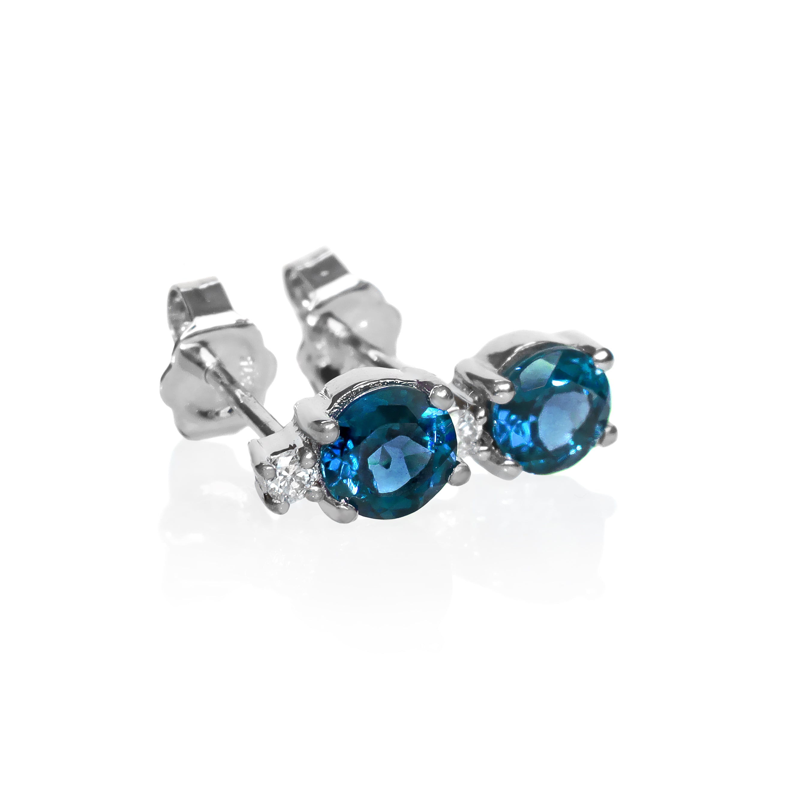 A product photo of a pair of white gold london blue topaz earrings sitting against a white background. The simple circle-cut stones are complimented by a single white diamond atop each stone, encased in white gold.
