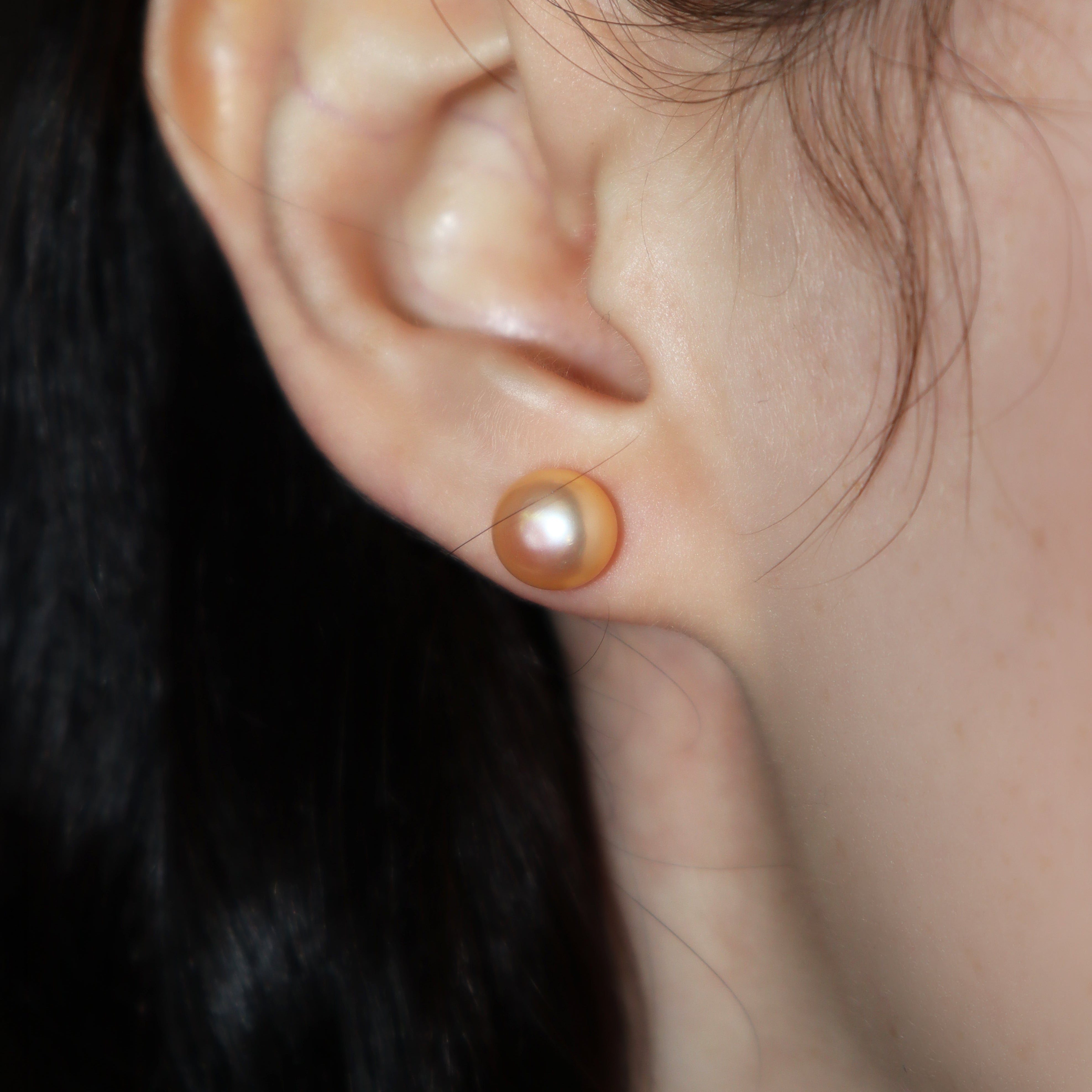 A product photo of simple peach-coloured pearl yellow gold earring studs photographed on a model's earlobe.