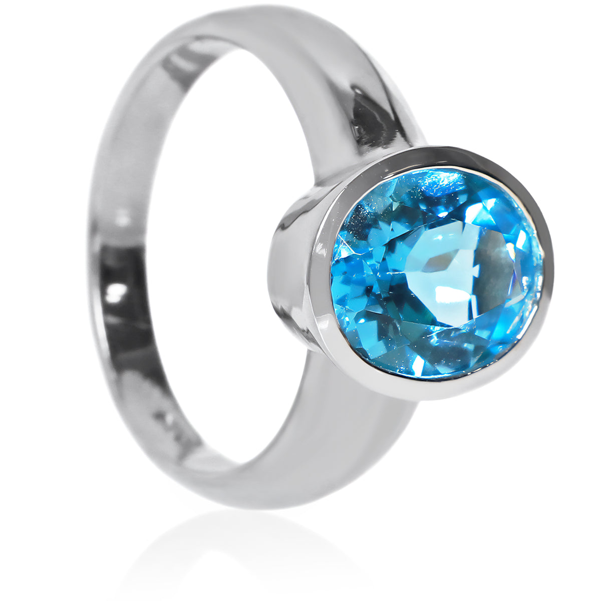 A product photo of a silver ring with a large bezel-set topaz centre stone sitting on a white background. The silver band is simple and smooth, connecting on either side of a vertically-oriented oval-cut topaz stone surrounded by a solid frame of silver. The topaz jewel is almost the colour of a tropical sea, reflecting bright blue light across its multi-faceted surface.