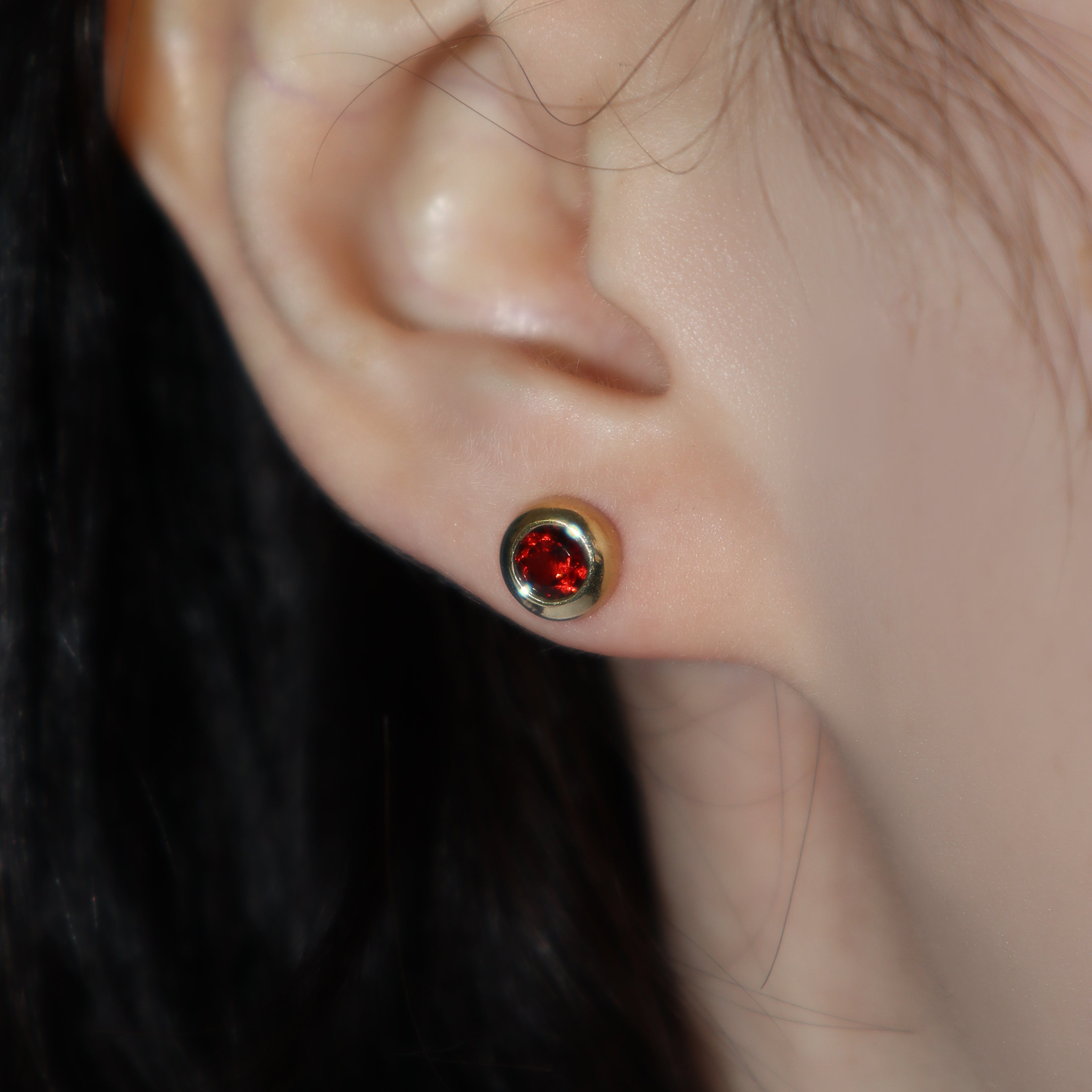 A product photo of 4mm Round Bezel-Set Garnet Earring Studs in 9k Yellow Gold photographed on a model's earlobe. The 2 garnet stones measure 4mm across and are deep red, reflecting sanguine hues across their multi-faceted edges, framed in a thick layer of yellow gold.