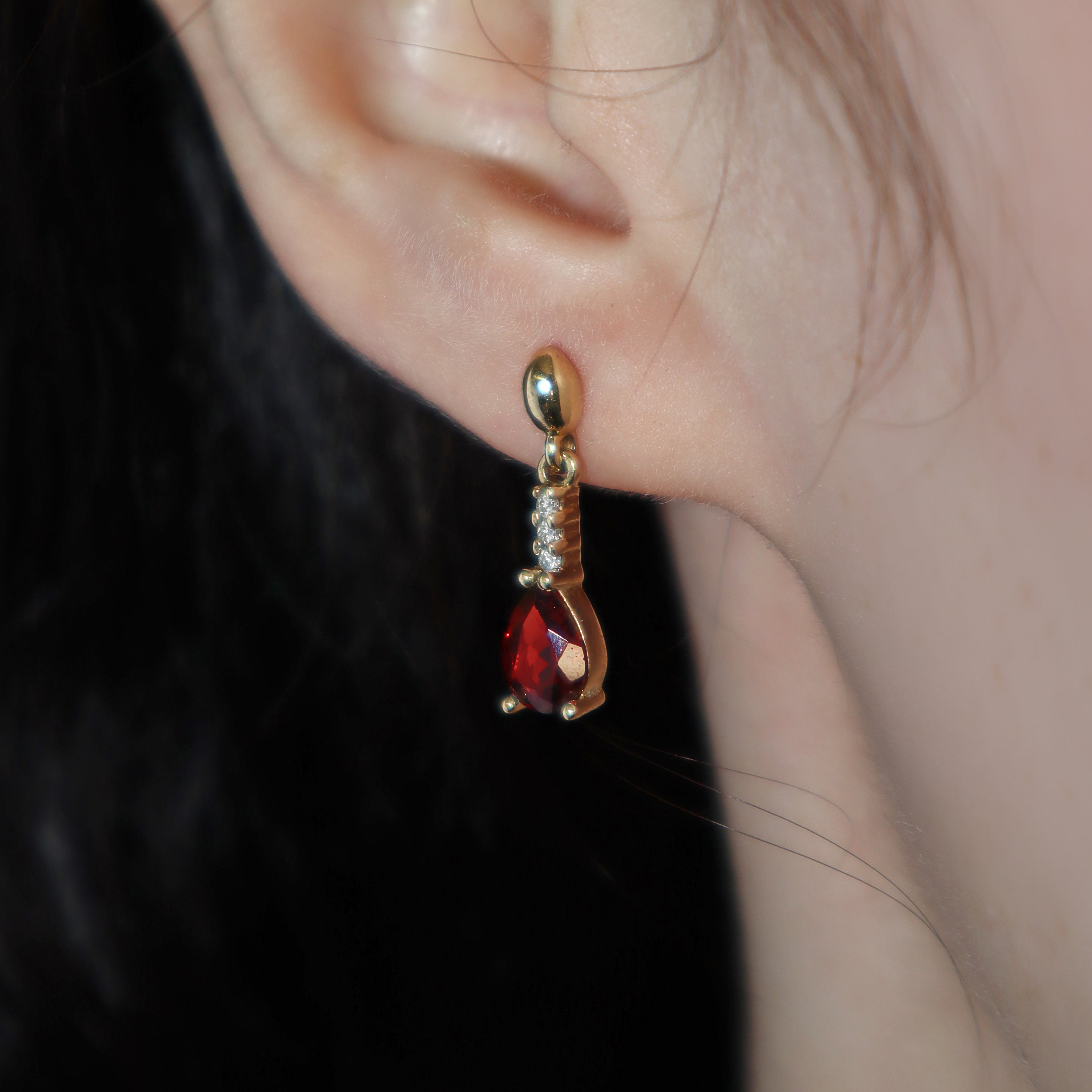 A product photo of 7x5mm Pear Garnet & Diamond Earrings in 9k Yellow Gold photographed on a model's earlobe. A golden strip connects the garnets to the studs, each strip adorned with 3 diamonds each. The garnets reflect sanguine hues across their multi-faceted edges.