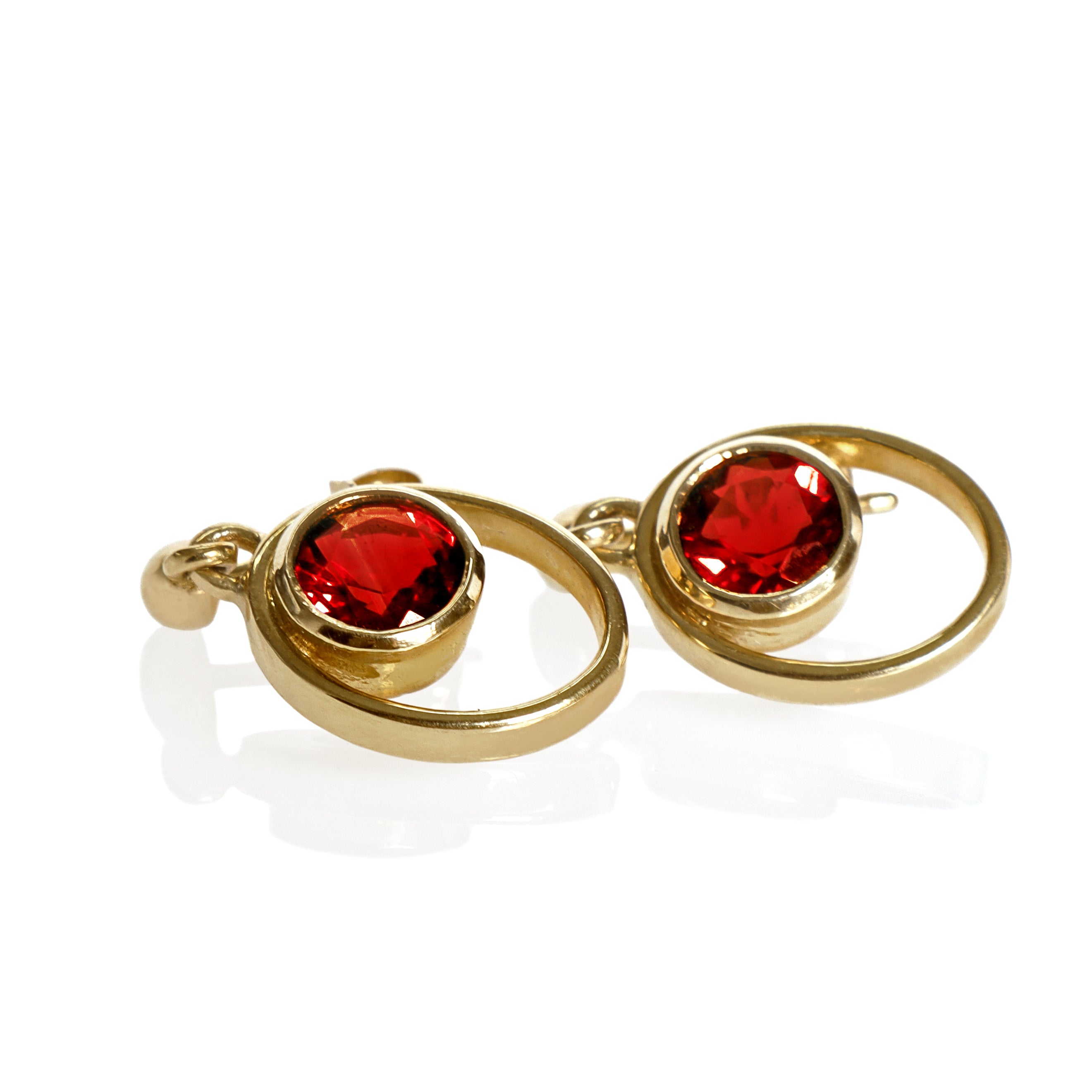 A product photo of 5.5mm Round Bezel-set Garnet Earring Studs in 9k Yellow Gold sitting on a plain white background. The bezel-set stones are nestled at the top of golden oval loops of similar thickness to the bezel frames. The garnets reflect sanguine hues across their multi-faceted edges.