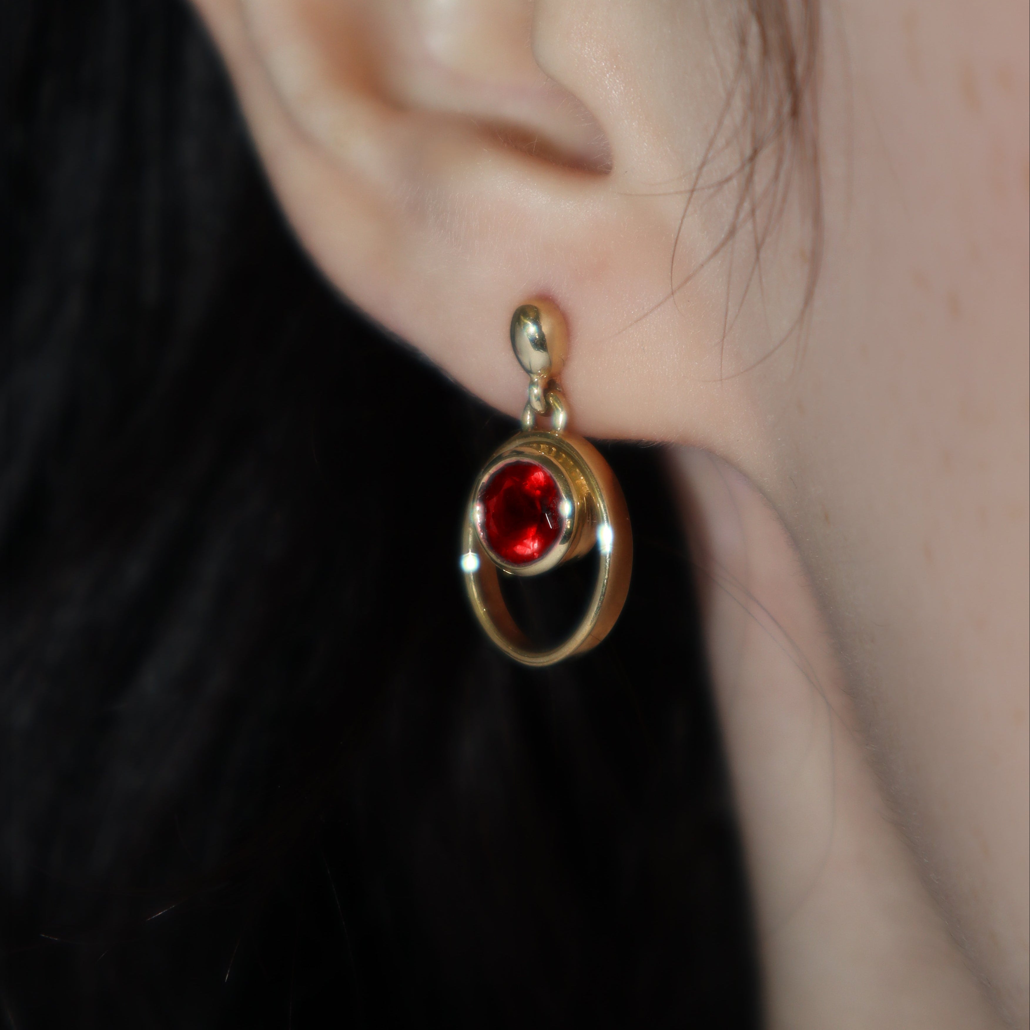 A product photo of 5.5mm Round Bezel-set Garnet Earring Studs in 9k Yellow Gold photographed on a model's earlobe. The bezel-set stones are nestled at the top of golden oval loops of similar thickness to the bezel frames. The garnets reflect sanguine hues across their multi-faceted edges.