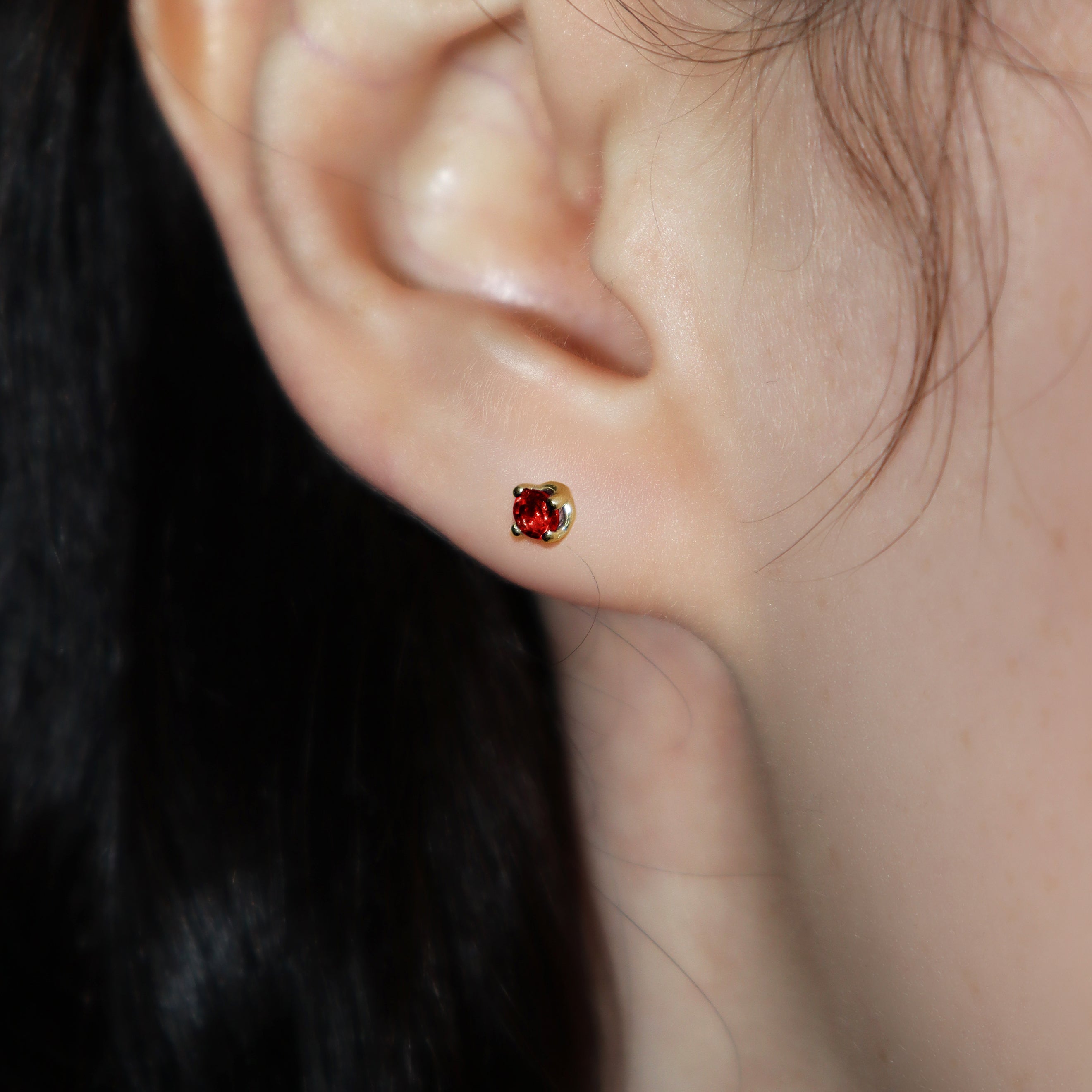 A product photo of 3.5mm Round Garnet Earring Studs in 9k Yellow Gold sitting on a plain white background. The stones are held in place by 4 delicate golden claws arranged in a flower-like pattern where they meet the stud. The garnets reflect sanguine hues across their multi-faceted edges.