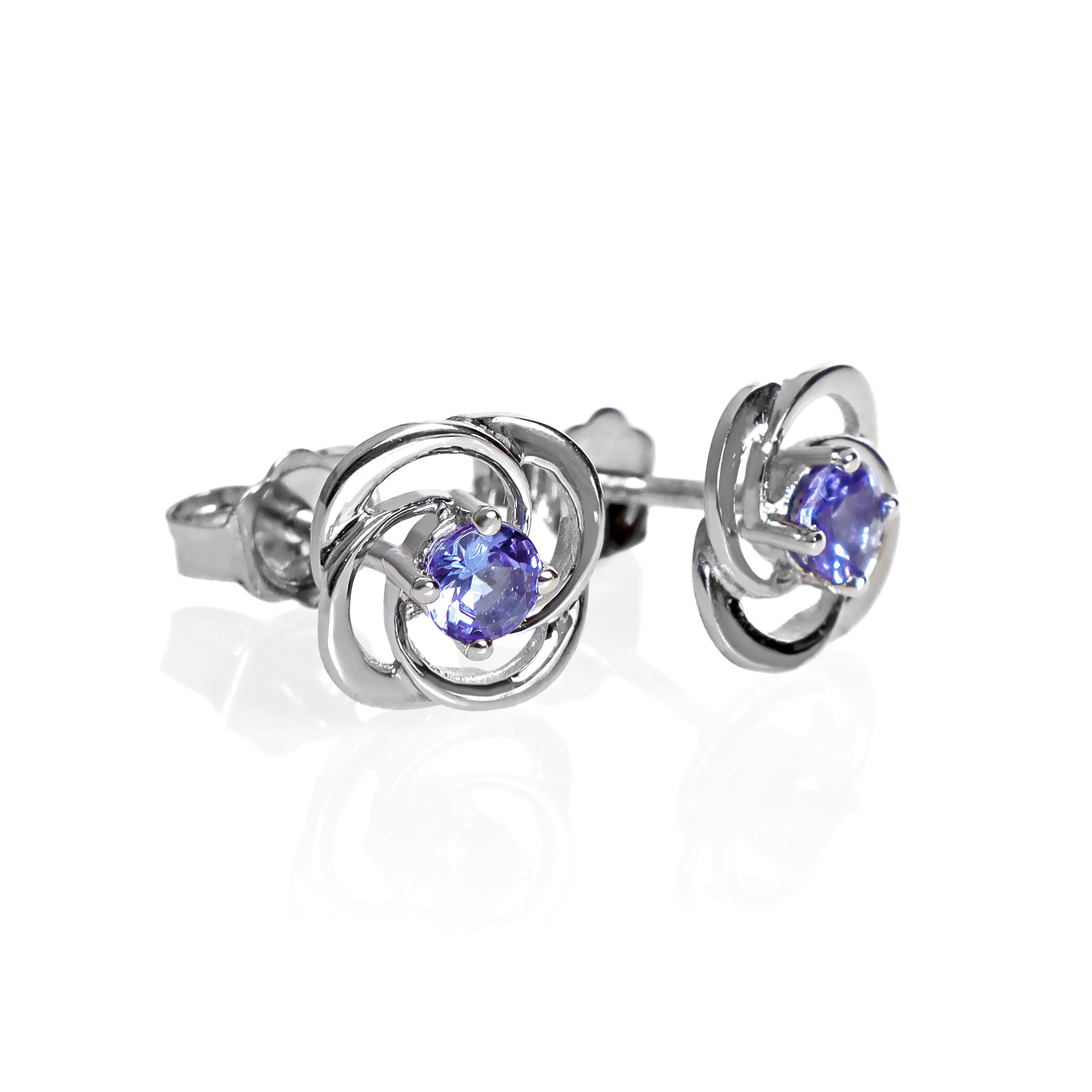 A product photo of white gold stud tanzanite earrings sitting on a white background. The small stones are framed by ornate, flowery white gold backs. The deep blue tanzanite stones reflect violet blue and indigo light from their many edges.