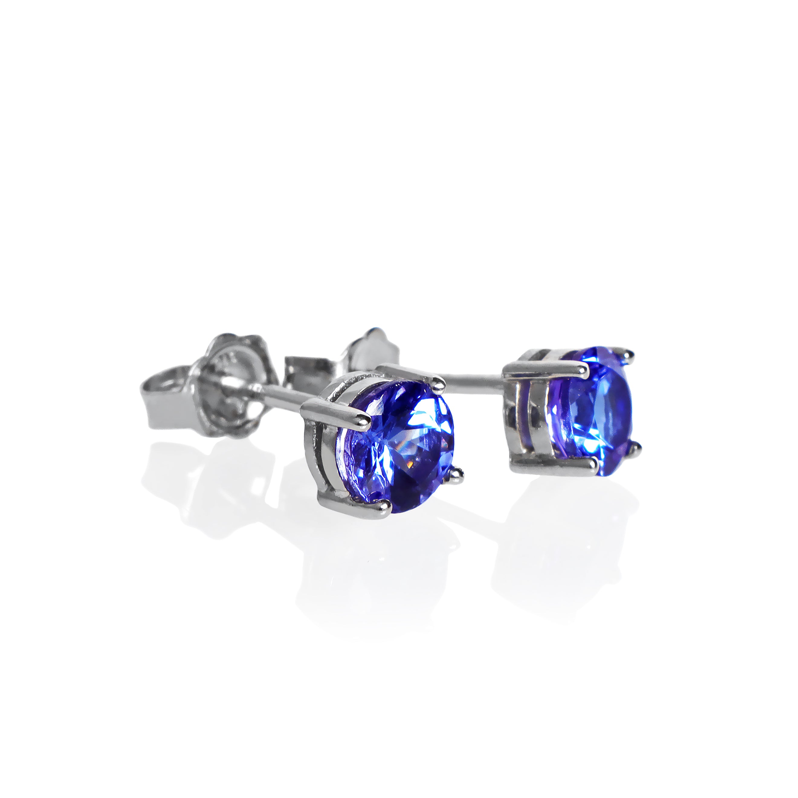 A product photo of white gold tanzanite stud earrings sitting on a white background. The round stones are held in place by 4 claws each, and reflect blue and indigo light from their many edges.