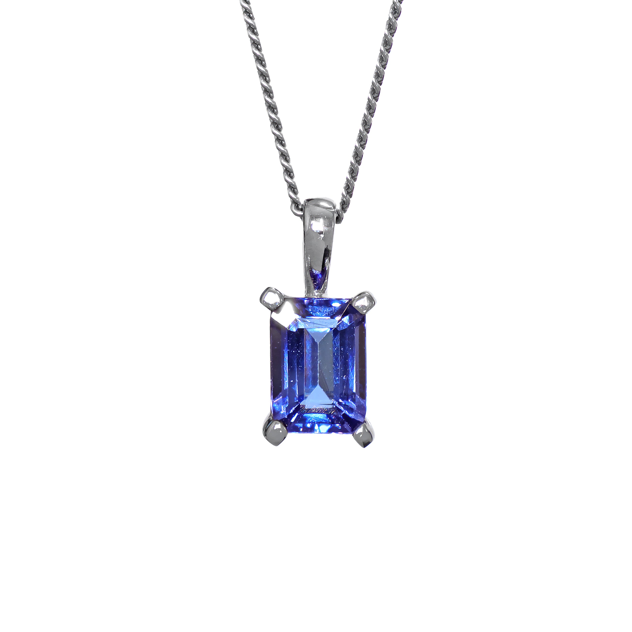 A product photo of a white gold tanzanite pendant suspended by a chain against a white background. The emerald-cut tanzanite stone is held in place by 4 golden claws, and reflects violet blue and indigo colours from its edges.