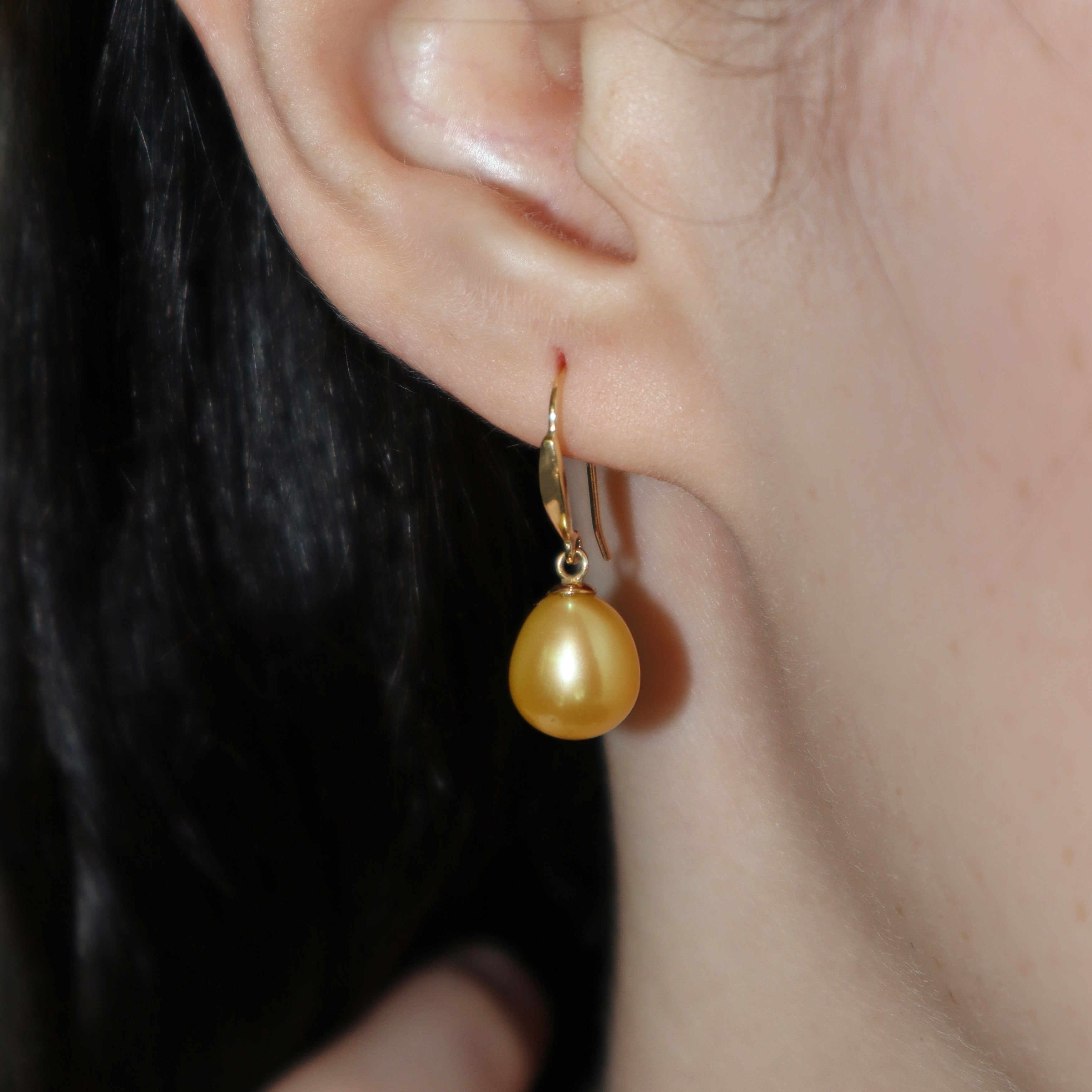 A product photo of a simple pair of yellow gold shepherd's hook earrings suspending two golden pearls photographed on a model's earlobe.