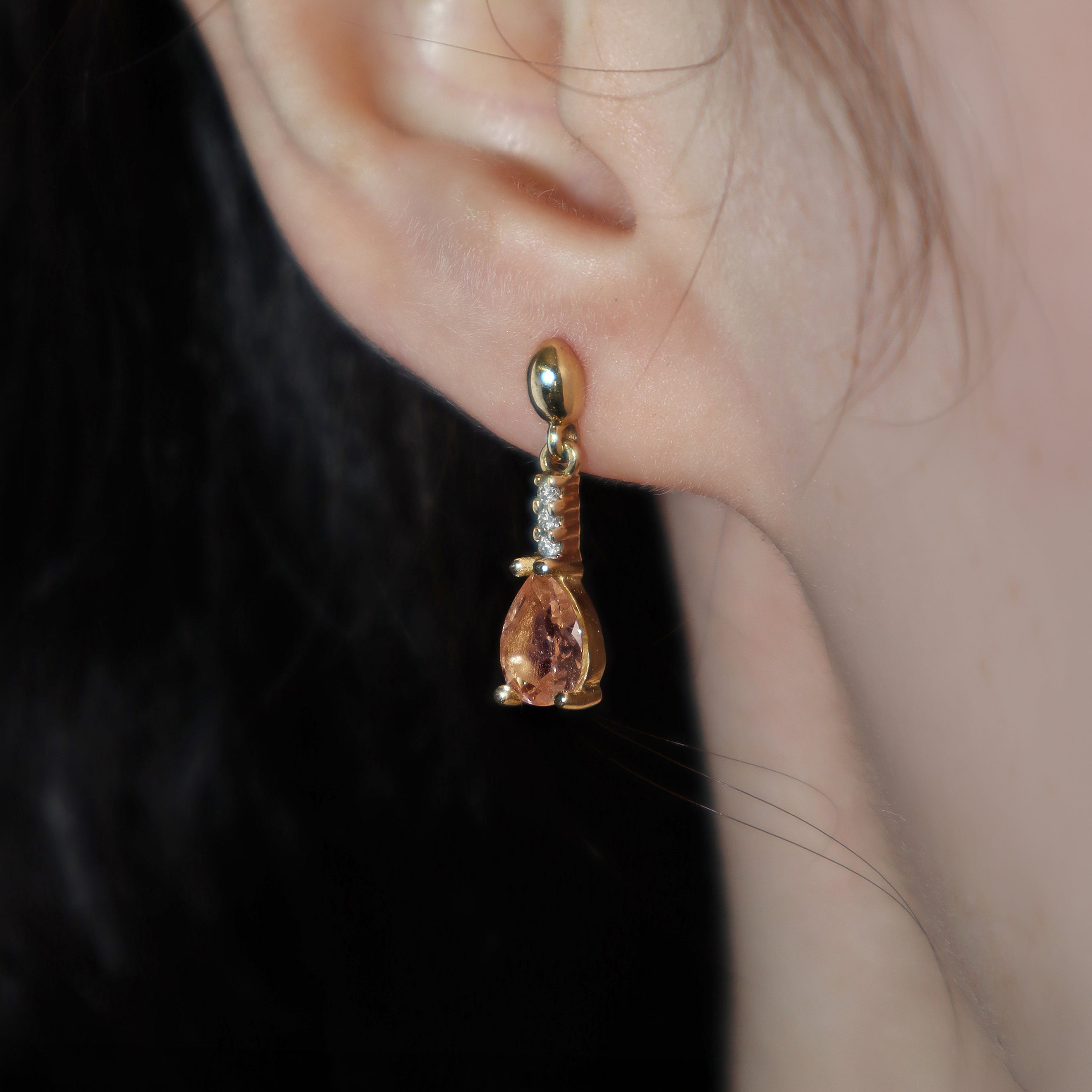 A product photo of 7x5mm Pear Morganite & Diamond Earrings in 9k Yellow Gold sitting on a plain white background. A golden strip connects the morganites to the studs, each strip adorned with 3 diamonds each. The morganites reflect light pinky hues across their multi-faceted edges.