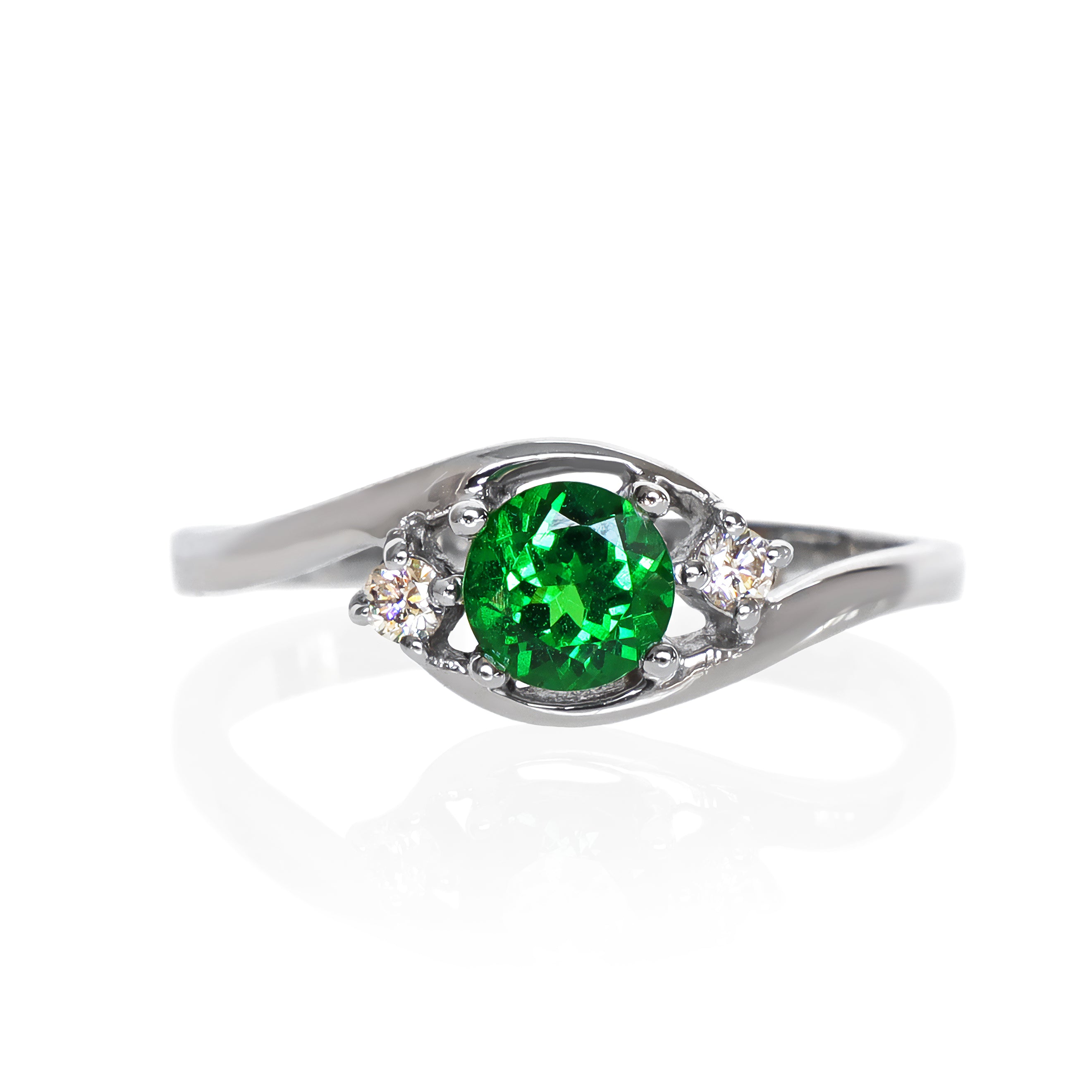 A product photo of a mystical tsavorite and diamond fantasy ring in solid 9k white gold on a white background. The deep, verdant-green tsavorite is held in place by 4 claws and reflects light off of its many facets, while the diamonds on either side are held by 3 each. The smooth white gold band curves elegantly, meeting at the top and bottom of the tsavorite and diamond arrangement. The green gemstone colour be a good emerald substitute.