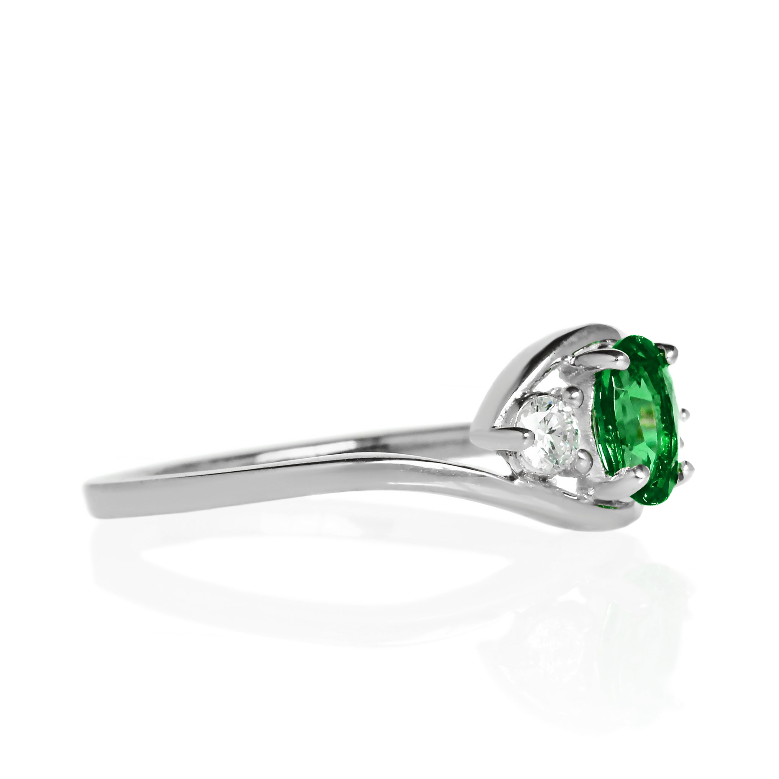 A product photo of a mystical tsavorite and diamond fantasy ring in solid 9k white gold on a white background. The deep, verdant-green tsavorite is held in place by 4 claws and reflects light off of its many facets, while the diamonds on either side are held by 3 each. The smooth white gold band curves elegantly, meeting at the top and bottom of the tsavorite and diamond arrangement. The green gemstone colour would be a good emerald substitute.