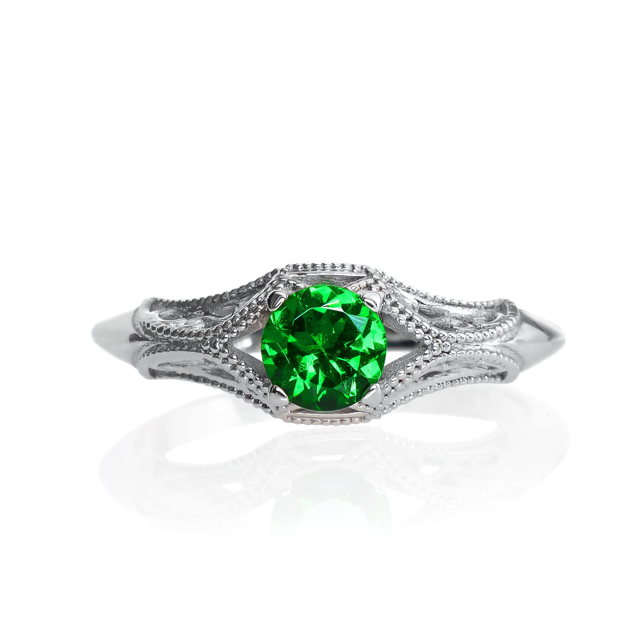 A product photo of an ornate, filigree-detailed white gold ring - reminiscent of an elvish fantasy ring with a deep forest green tsavorite centre stone. The band is detailed with delicate curves and patterns for about half of its length around the dazzling tsavorite stone, before smoothing out towards the back of the ring. The green gemstone colour would be a good emerald substitute.