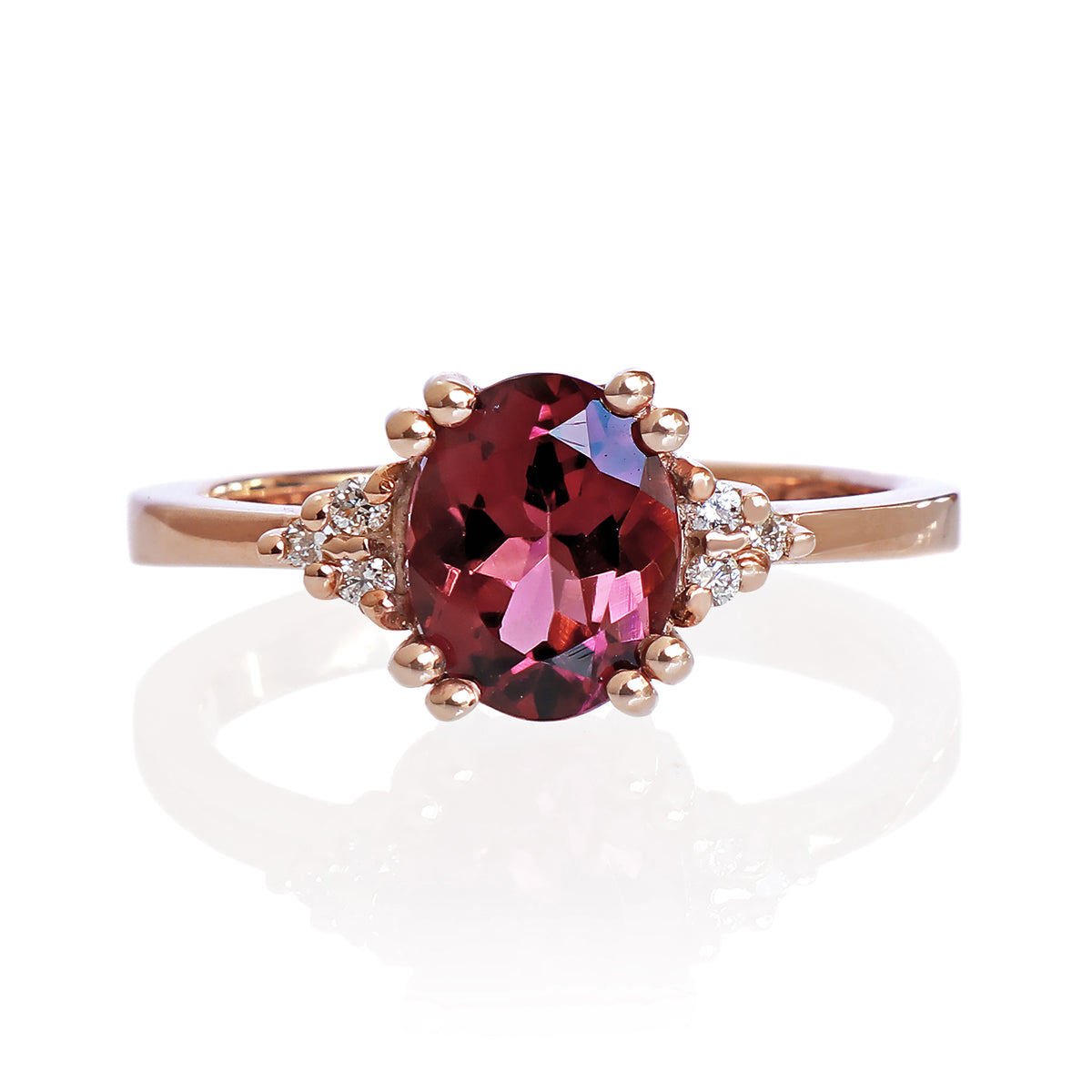 A product photo of a rose gold pink tourmaline and diamond trio ring sitting on a white background. The oval magenta pink tourmaline gemstone stands in stark contrast to the little clusters of three white diamond stones on either side.