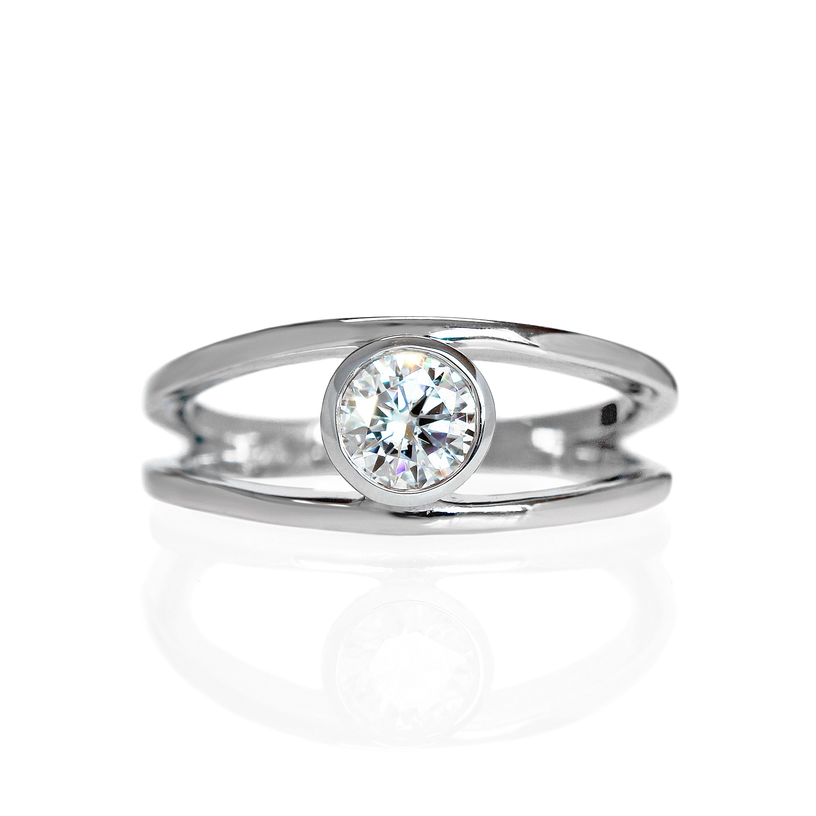A product photo of a split-band silver ring with a bezel-set white moissanite centre stone sitting on a white background. The silver band splits at the base of the ring, separating to meet at the top and bottom of the dazzling round-cut centre stone, framed in a thick layer of silver, holding it in place.