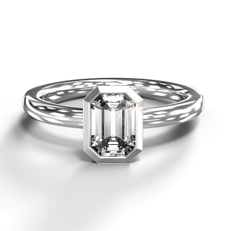 A turntable video of a 14 karat white gold lab diamond bezel solitaire engagement ring sitting in the sun on a white textured background. The brilliant, colourless 0.72ct emerald-cut diamond measures about 6.3mm lengthwise, and is held in place by a delicate golden-framed bezel setting.
