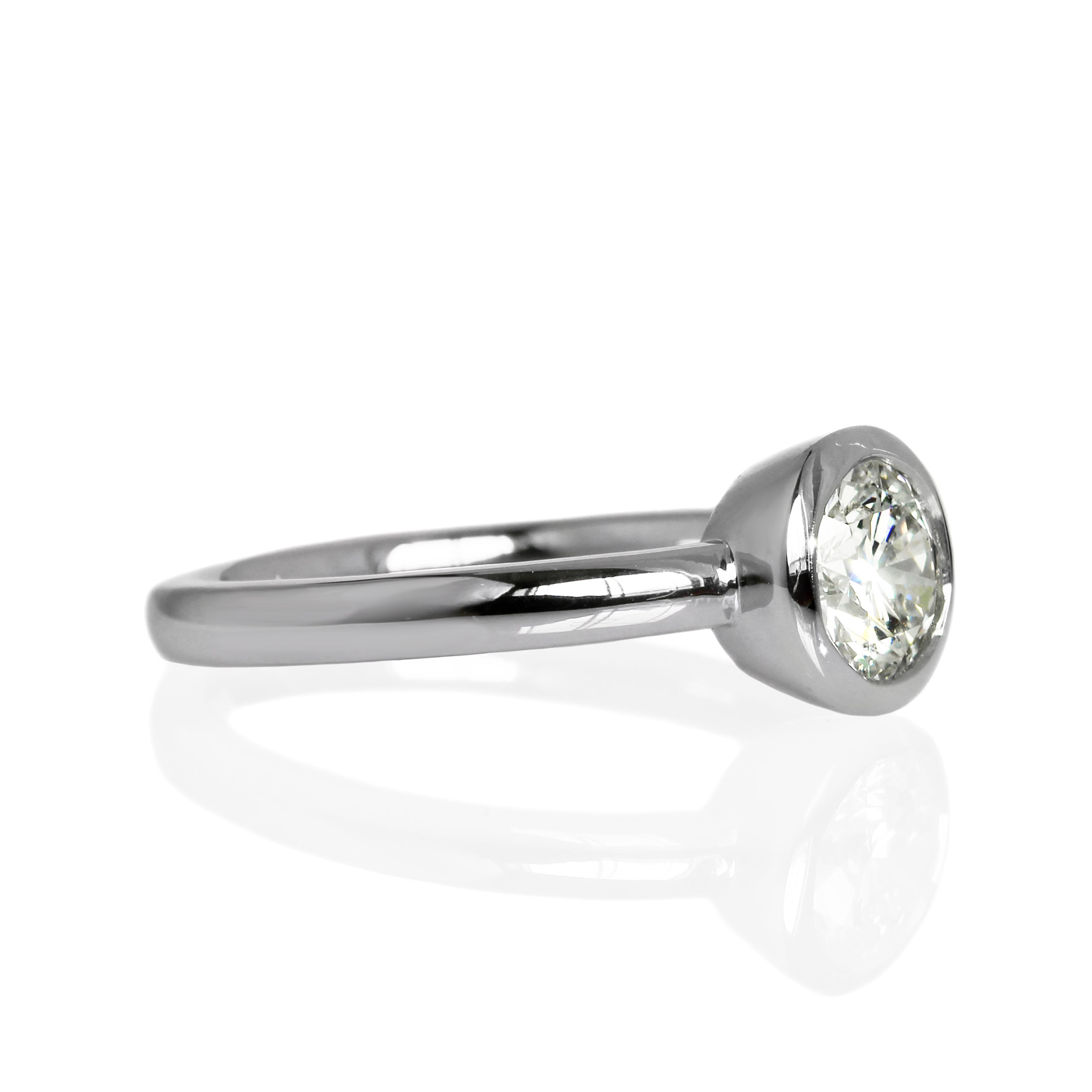 A product photo of a 14 karat white gold lab diamond solitaire engagement ring sitting in the sun on a white textured background. The brilliant, colourless 1 carat round-cut diamond measures 6.3mm lengthwise, and is held in place by a thick frame of white gold in a bezel setting.