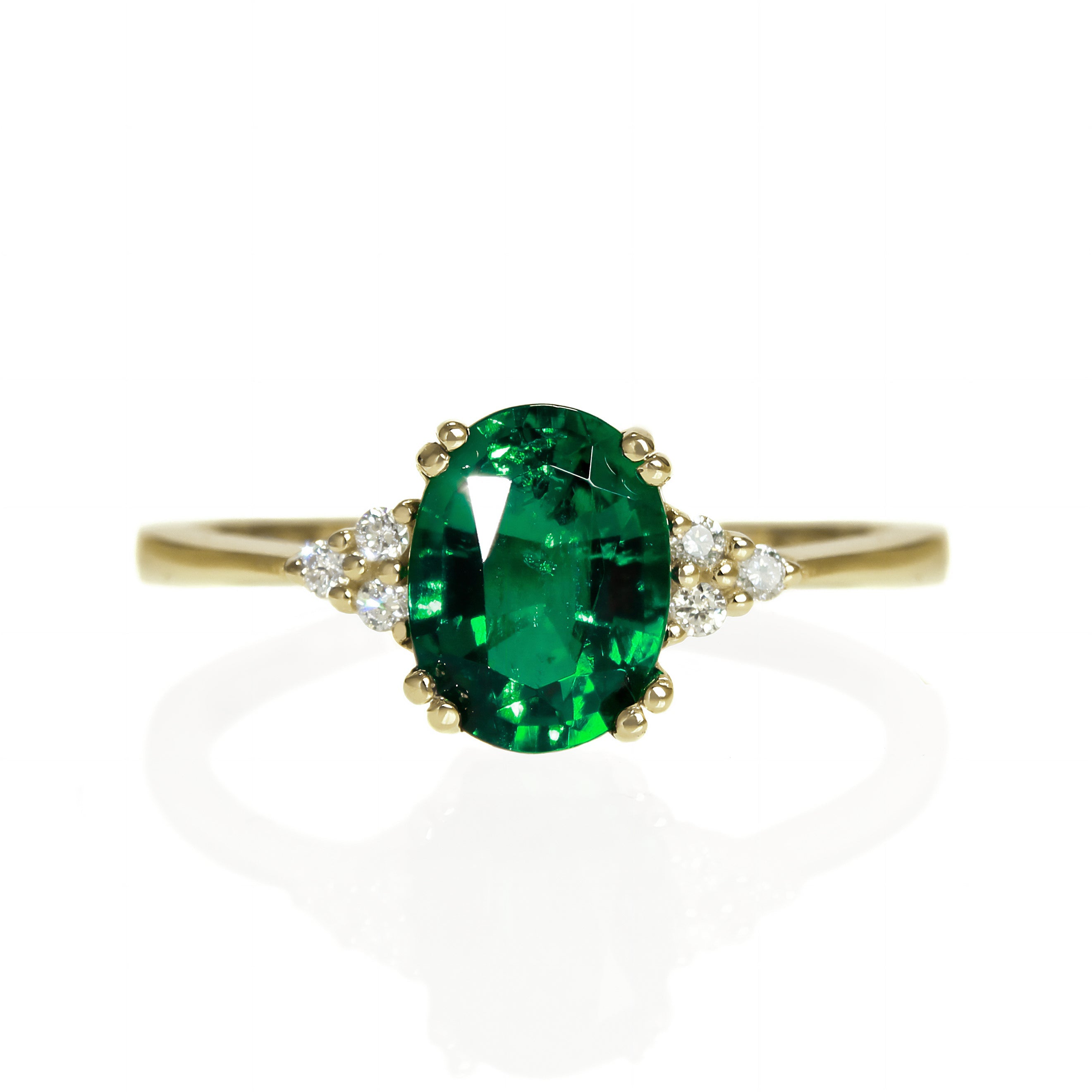 A product photo of a yellow gold lab grown emerald and lab grown diamond trio ring sitting on a white background. The oval, eyeclear May birthstone is a vibrant electric green, almost the same colour as a tsavorite garnet, and its colouring stands in stark contrast to the little clusters of three classic white lab diamond stones on either side.