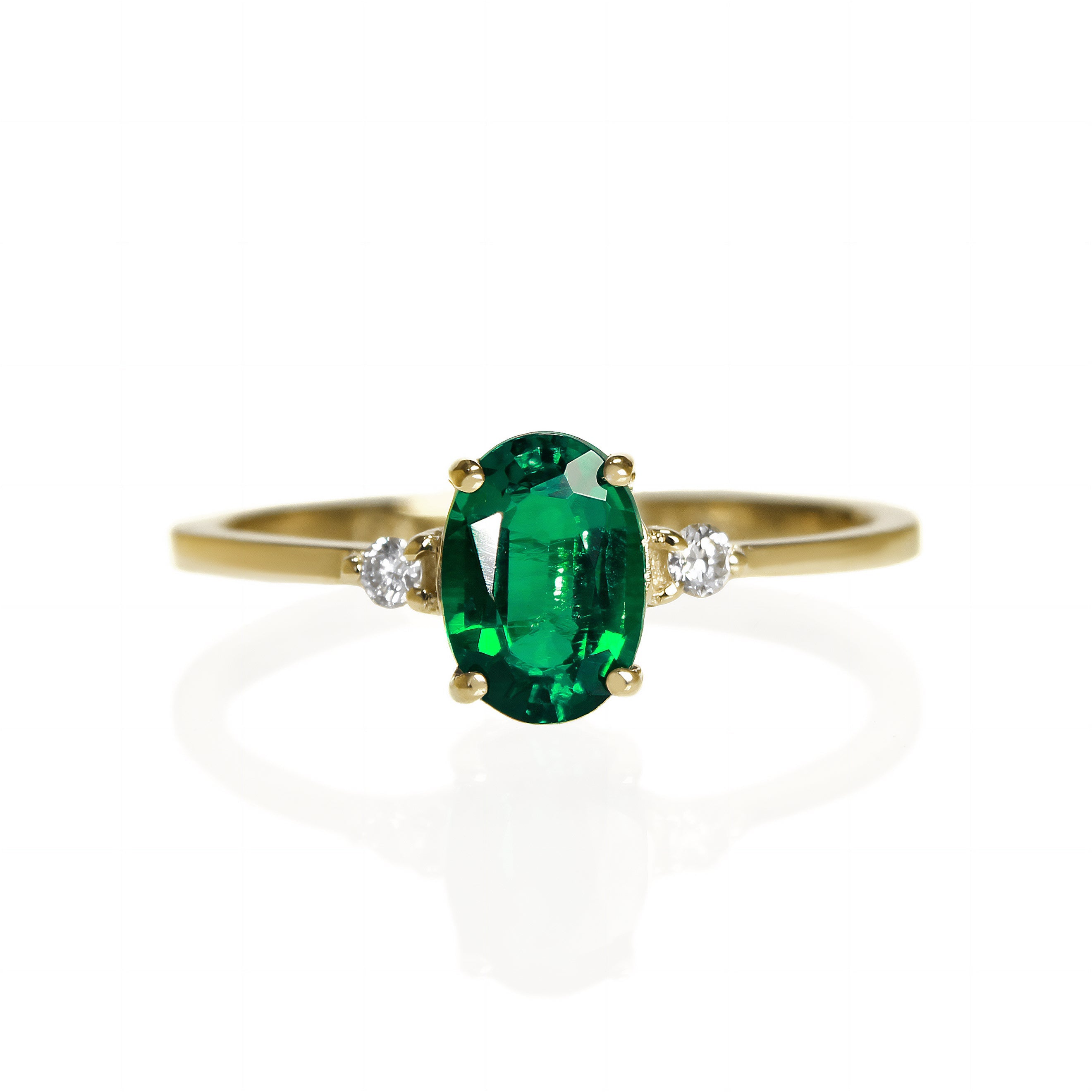 A product photo of a yellow gold lab emerald ring sitting against a white background. The yellow gold band is plain and smooth, and the centre oval-cut electric green May birthstone jewel is framed by a single white lab grown diamond on either side.