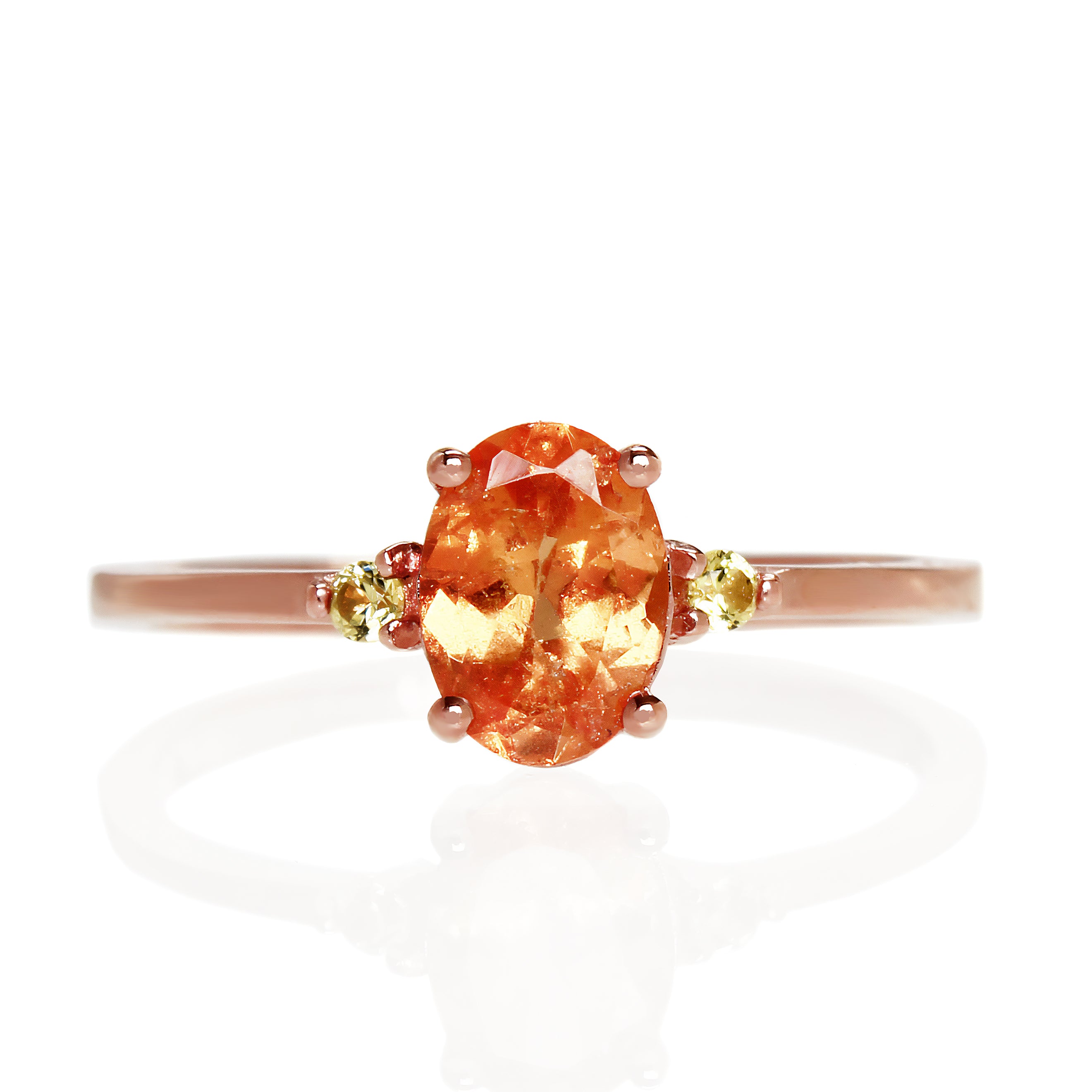 A product photo of a rose gold mandarin garnet ring sitting against a white background. The rose gold band is plain and smooth, and the centre oval-cut light bright orange centre stone is framed by a single yellow sapphire jewel on either side.