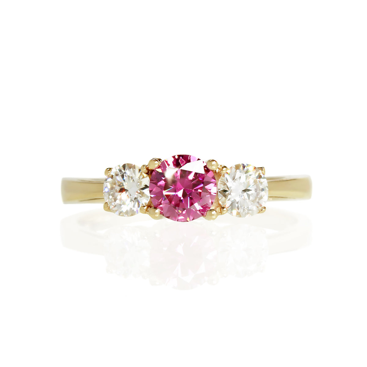 A product photo of a classic trilogy-style engagement ring set in solid 9 karat yellow gold on a plain white background. The ring consists of two smaller brilliant moissanite stones. each 4mm, sitting on either side of the 5mm fancy pink coloured moissanite centre stone - all held in place by traditional basket setting.