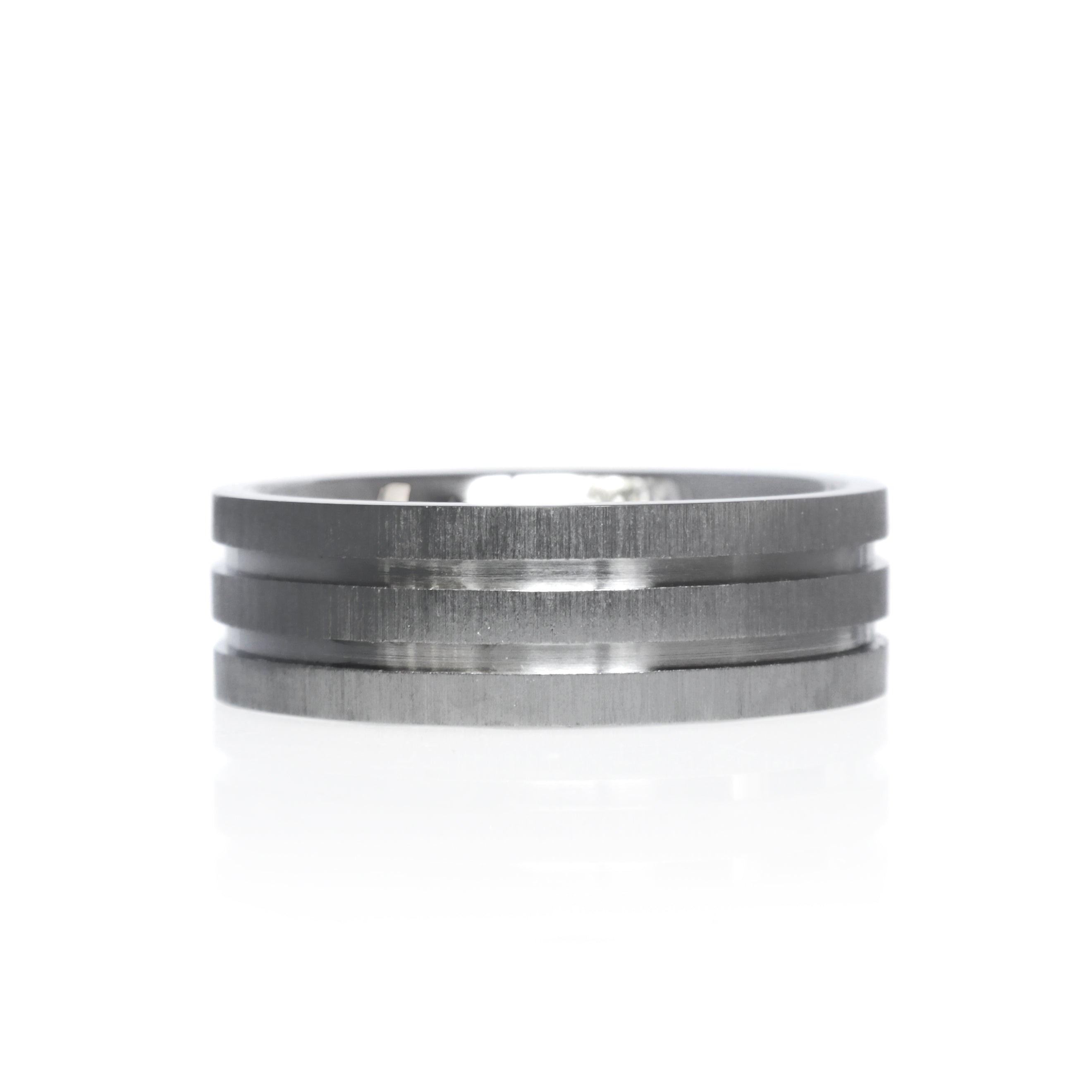 A product image of a 7mm wide ring against a plain white background. The ring is crafted from titanium with a brushed finish across the center band. Two grooves are etched into the titanium, creating a double-grooved design that runs horizontally around the ring. The edges of the ring are polished, providing a subtle contrast to the brushed center.