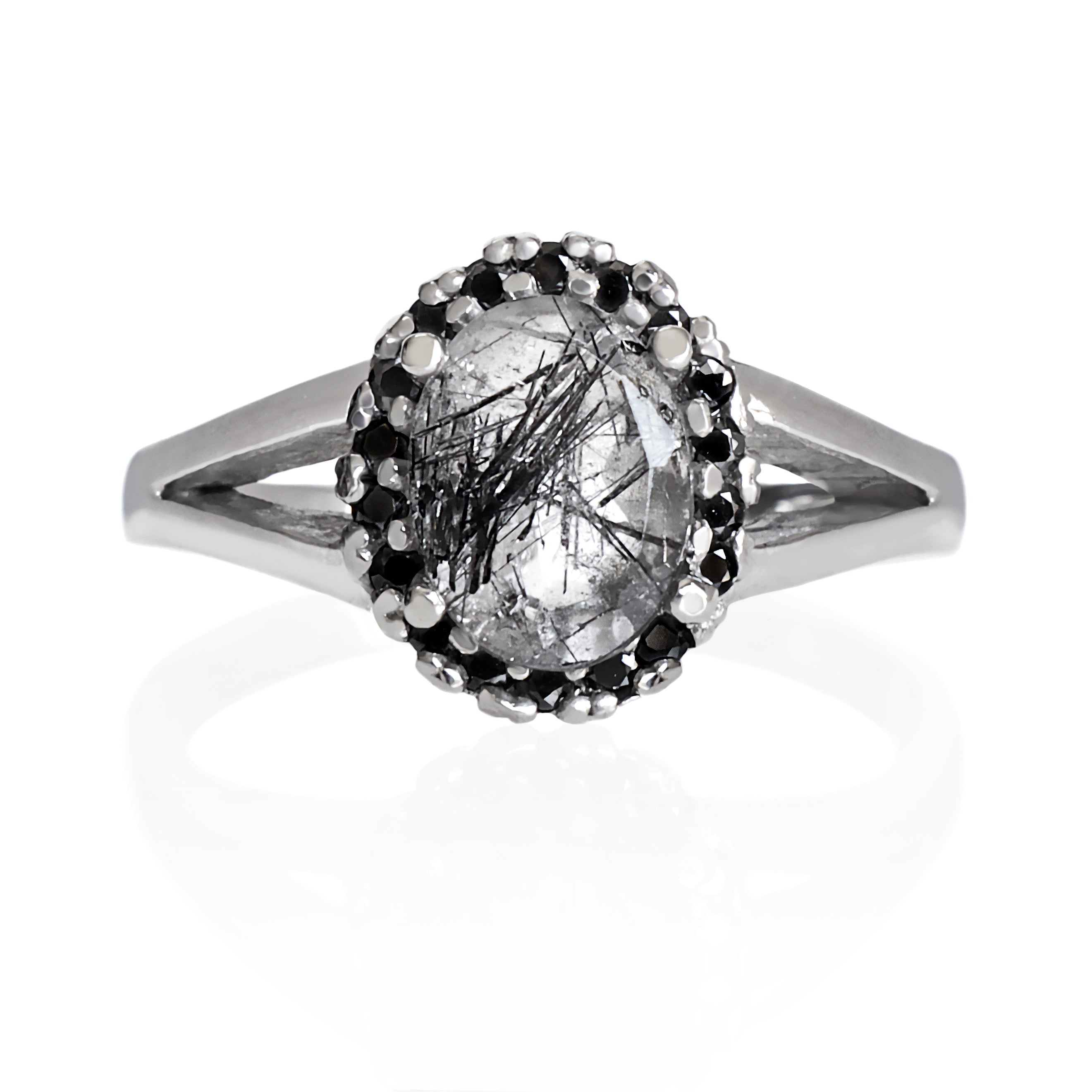 A product photo of a tourmalated quartz ring with a lavish black moissanite halo in solid sterling silver on a white background. The smooth band splits in two halfway along its length on either side, meeting the halo and tourmalated quartz centre stone in the middle. The impressively large quartz is held in place by 4 claws and is heavily included, with black striations and and strikes snaking throughout the black and white jewel.