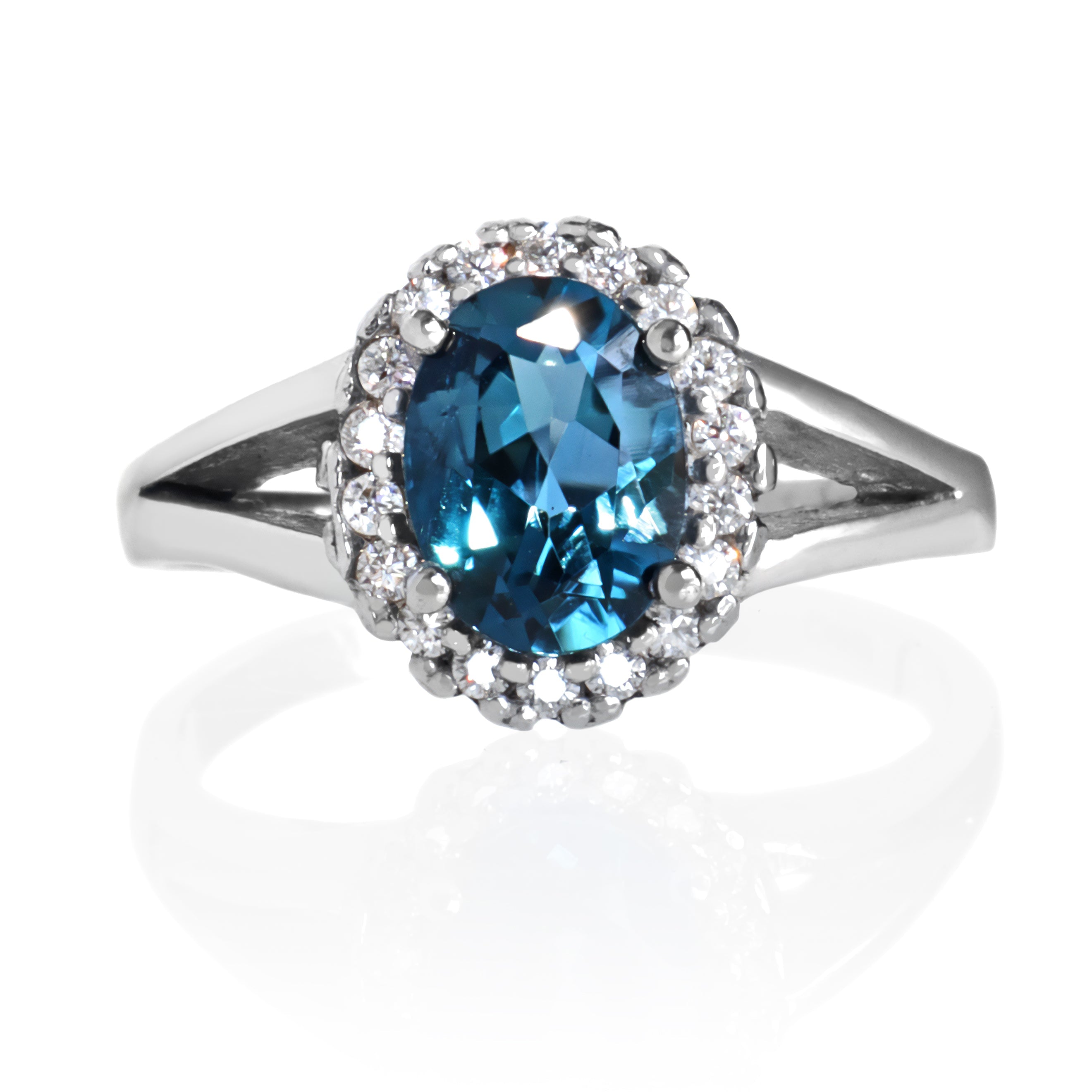 A product photo of a London Blue Topaz ring with a lavish moissanite halo in solid sterling silver on a white background. The smooth band splits in two halfway along its length on either side, meeting the halo and topaz in the centre. The impressively large ocean bluel stone is held in place by 4 claws.