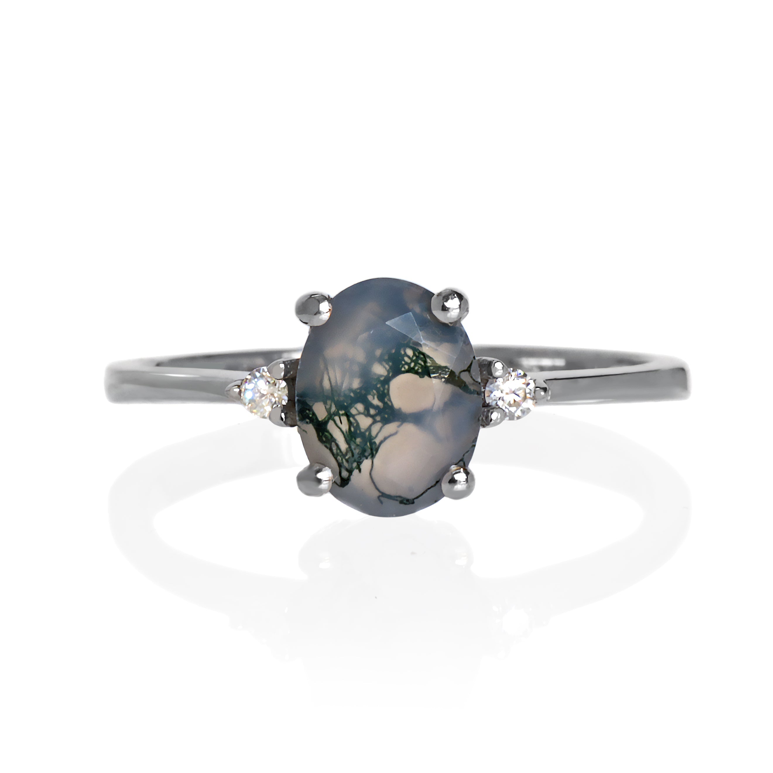 A product photo of a silver moss agate and moissanite ring sitting against a white background. The solid sterling silver band is plain and smooth, and the central oval-cut moss agate stone is framed by a single white moissanite jewel on either side.