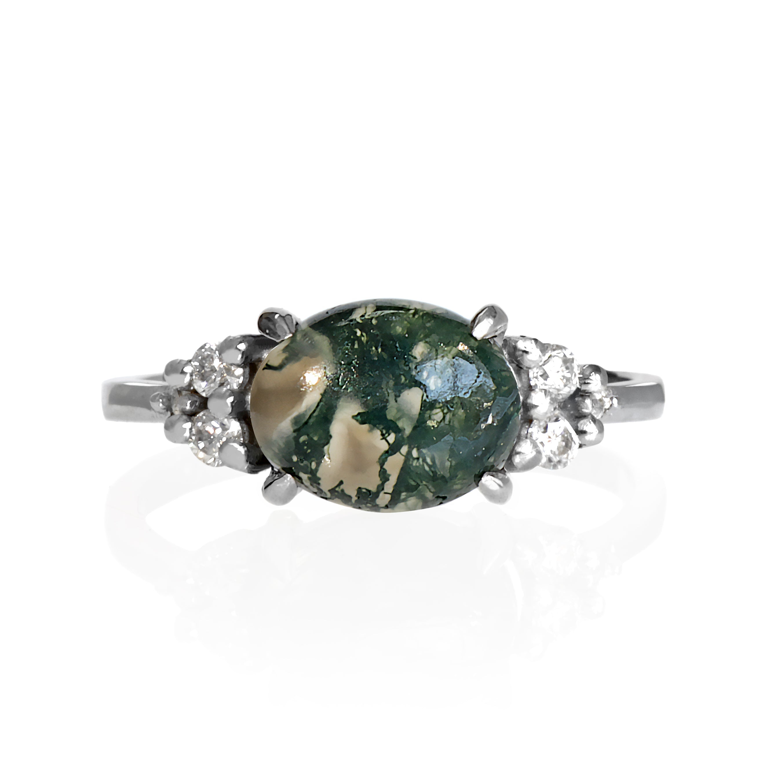 A product photo of a solid sterling silver moss agate and moissanite trio cluster ring sitting on a white background. The large oval moss agate gemstone stands in stark contrast to the little clusters of three white sparkling moissanite stones on either side.