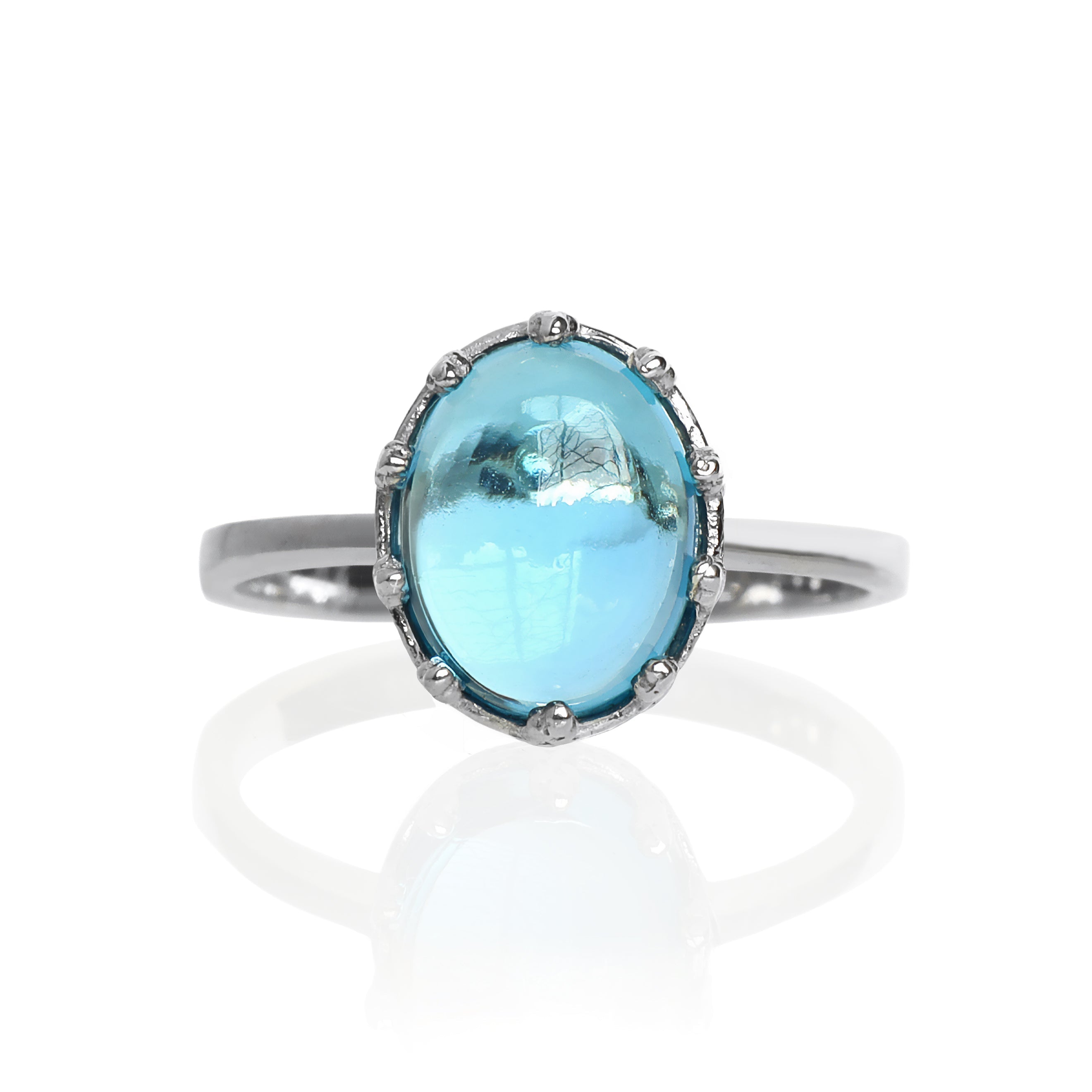 A product photo of a 925 sterling silver blue topaz birthstone ring sitting on a white background. The swiss blue topaz stoe is impressively large, with the cabochon cut allowing the viewer to look into the stone like a crystal ball. The 10x8mm oval stone is encased in ornate silver detailing, almost like a royal crown.