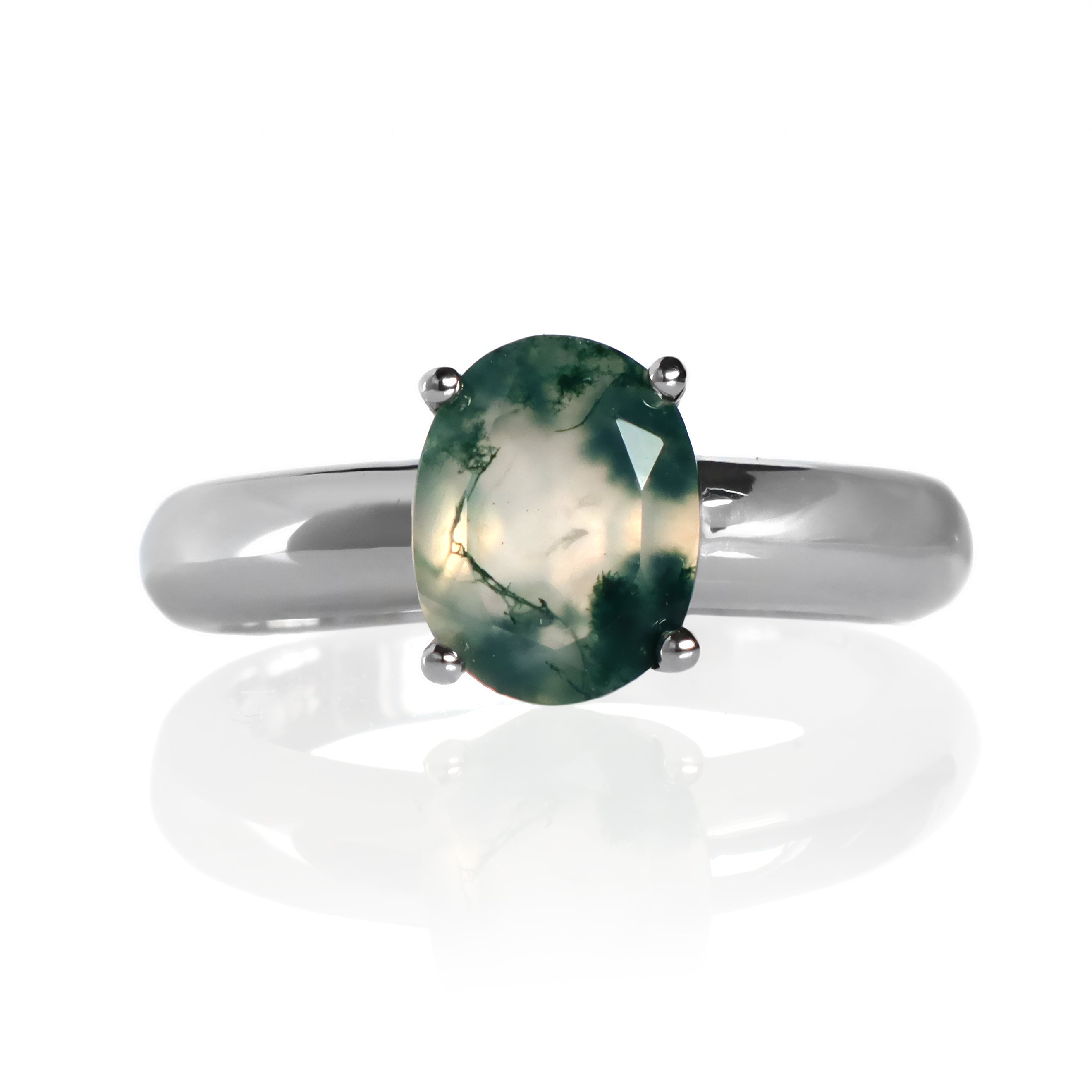 A product photo of a 9k white gold moss agate ring sitting on a white background. The ring features a prominent oval-cut moss agate gemstone as its centerpiece, held securely by four polished white gold prongs. The ring's band is a smooth, polished, and very thick 9k white gold, creating a classic and base setting for the gemstone. The moss agate displays a translucent, milky white to light green base with distinct deep green inclusions.