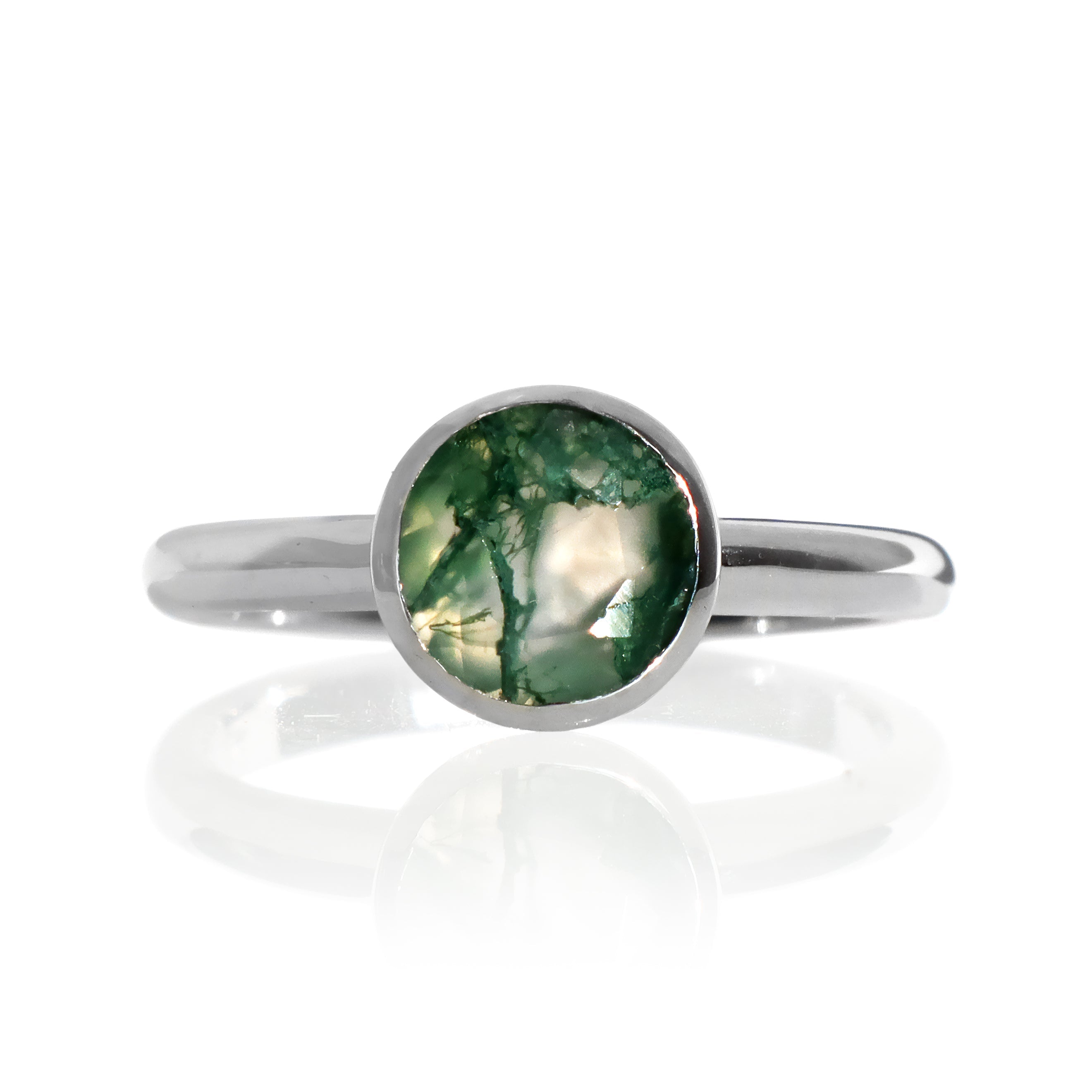 A product photo of a 9k white gold ring with a bezel-set moss agate centre stone sitting on a white background. The white gold band is simple and smooth, connecting on either side of a round-cut moss agate stone surrounded by a solid frame of gold.