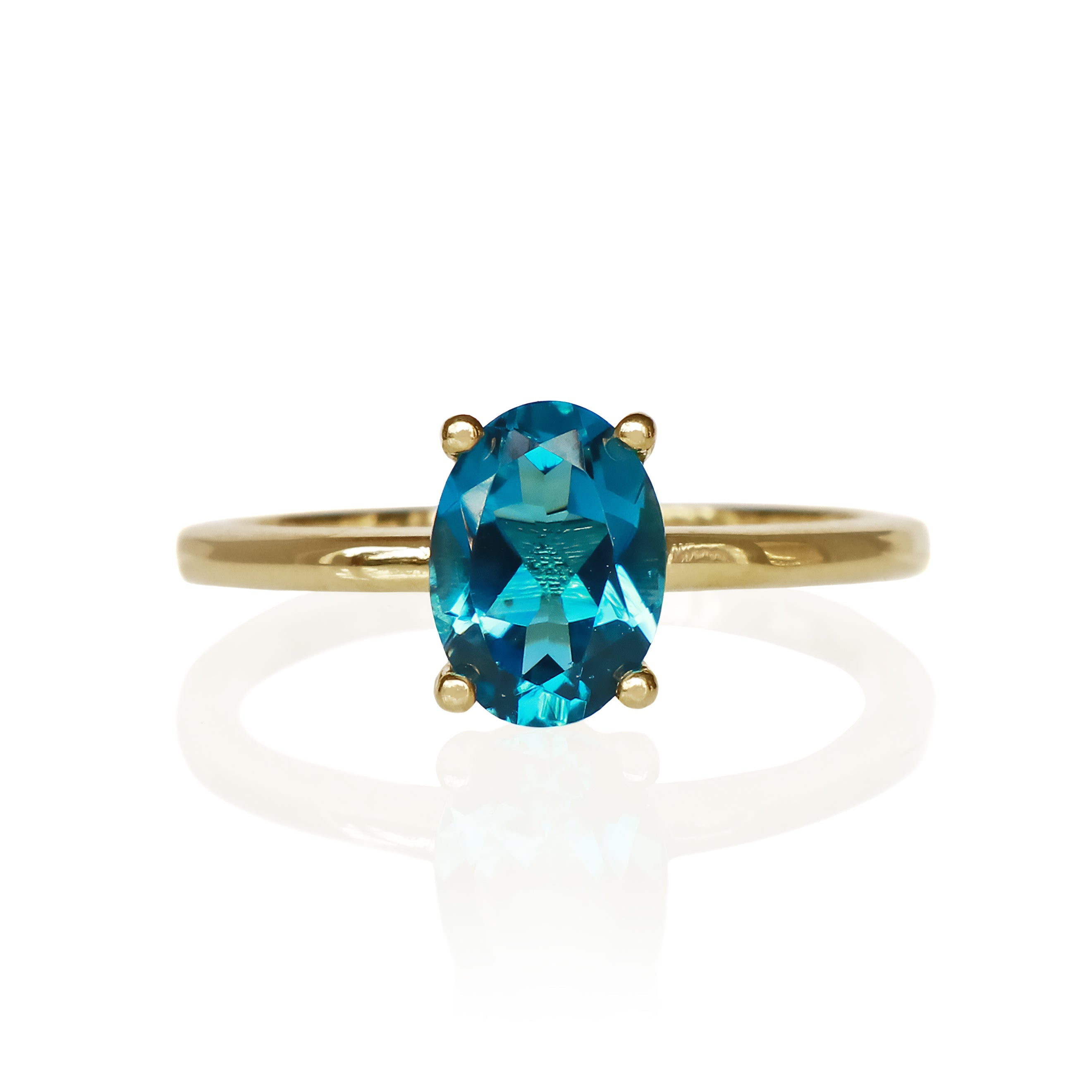 A product photo of a 9k yellow gold London blue topaz ring sitting on a white background. The ring features a remarkable 8x6mm oval-cut London blue topaz gemstone as its centerpiece, held by four polished yellow gold prongs. The London blue topaz displays a deep, rich teal-blue hue. The ring's band is a smooth, polished, and slender 9k yellow gold, offering a classic and elegant setting for the striking gemstone.