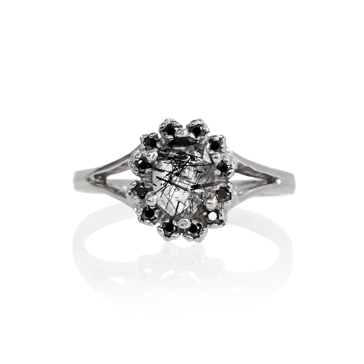 A product photo of a lavish floral moissanite halo tourmalated quartz ring in solid silver on a white background. The smooth band splits in two halfway along its length on either side, meeting the floral halo and tourmalated quartz in the centre. The tourmalated quartz centre stone is held in place by 4 claws and is lightly included, with natural black strikes snaking throughout the monochromatic jewel.