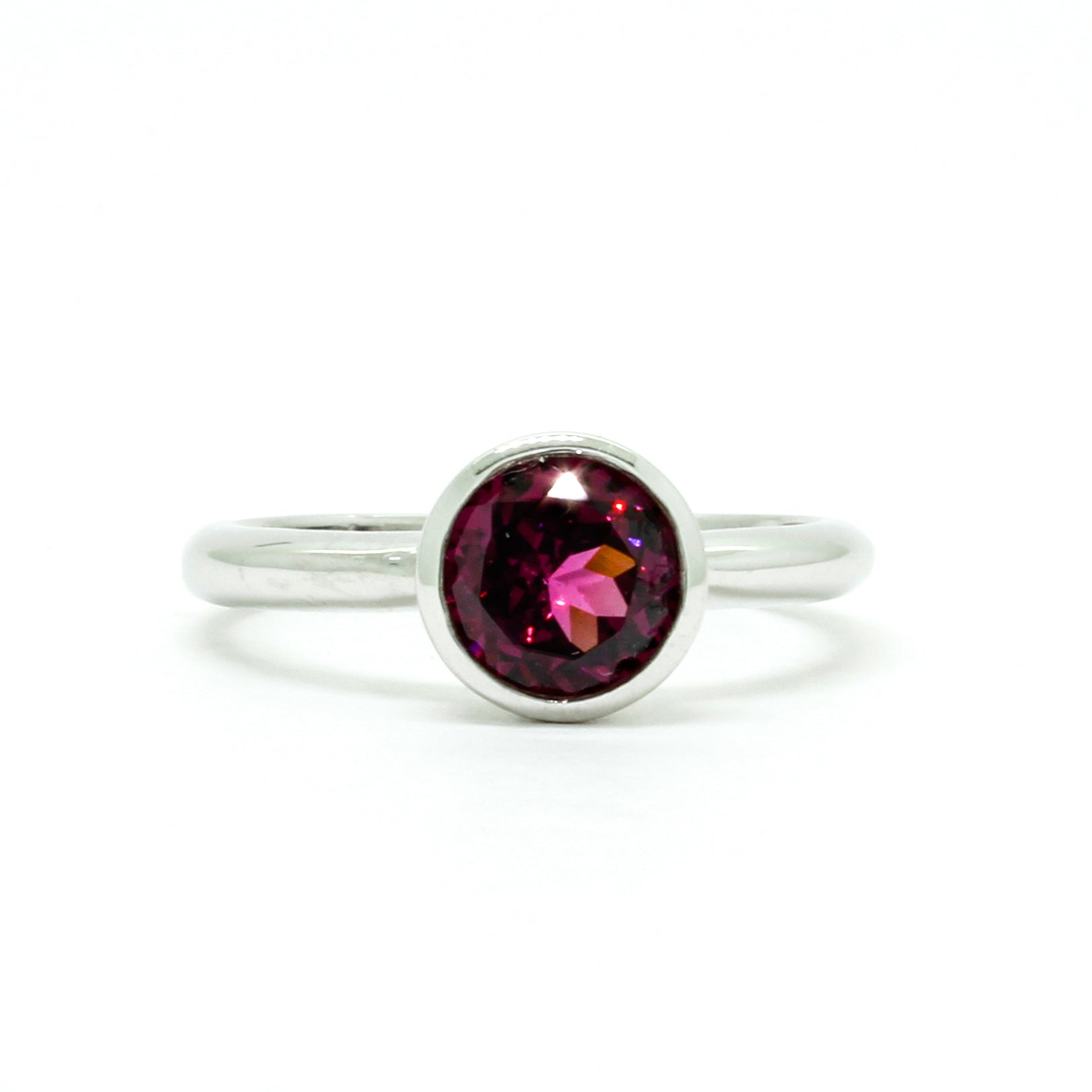 A product photo of a silver ring with a bezel-set rhodalite centre stone sitting on a white background. The silver band is simple and smooth, connecting on either side of a circle-cut rhodalite stone surrounded by a solid frame of silver. The rhodalite jewel is a deep velvet purple colour, reflecting a warm plum colours across its multi-faceted surface