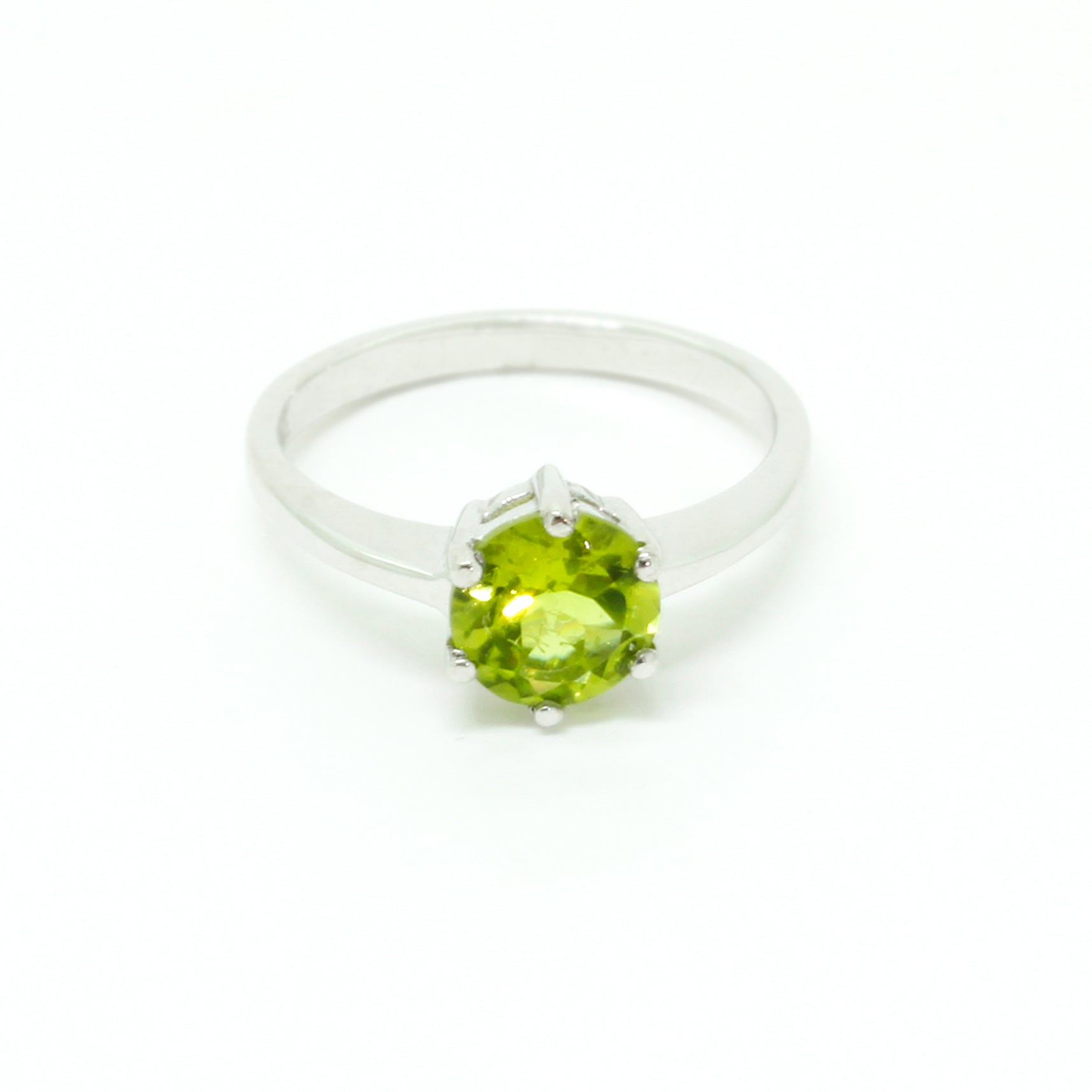 A product photo of a silver ring with a round-cut peridot centre stone sitting on a white background. The silver band is simple and smooth, connecting on either side of the circular peridot stone held in place by six silver claws. The peridot jewel is a shade of bright, vibrant green, reflecting chartreuse light across its multi-faceted surfaces.