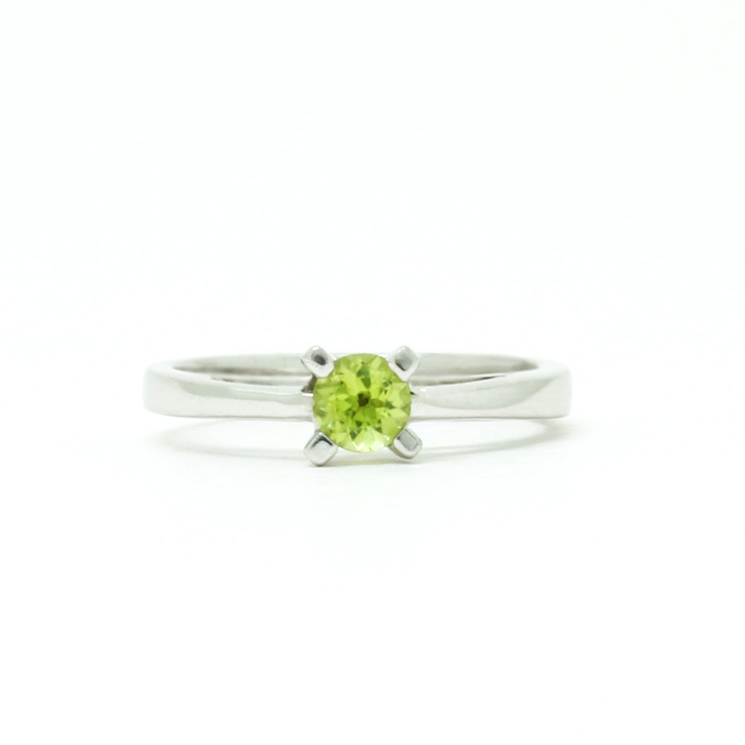 A product photo of a silver ring with a round-cut peridot centre stone sitting on a white background. The silver band is simple and smooth, connecting on either side of a small, roundl-cut peridot stone, held in place by four silver claws and a split band behind it. The peridot jewel is a shade of bright, vibrant green, reflecting chartreuse light across its multi-faceted surfaces.