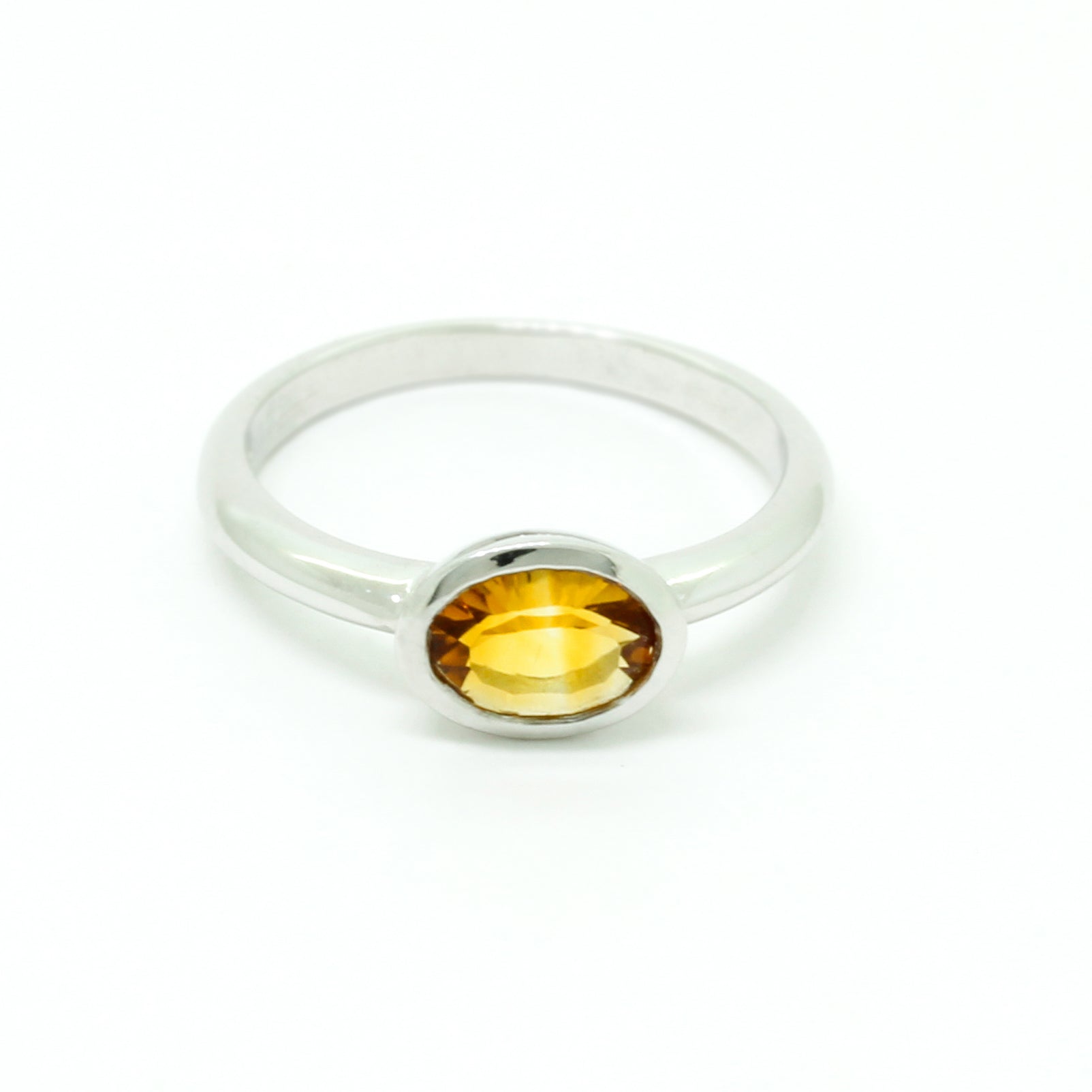 A product photo of a silver ring with a bezel-set citrine centre stone sitting on a white background. The silver band is simple and smooth, connecting on either side of a horizontally-oriented oval-cut citrine stone surrounded by a solid frame of silver. The citrine jewel is almost honey-coloured, reflecting a warm orangey yellow colour across its multi-faceted surface.