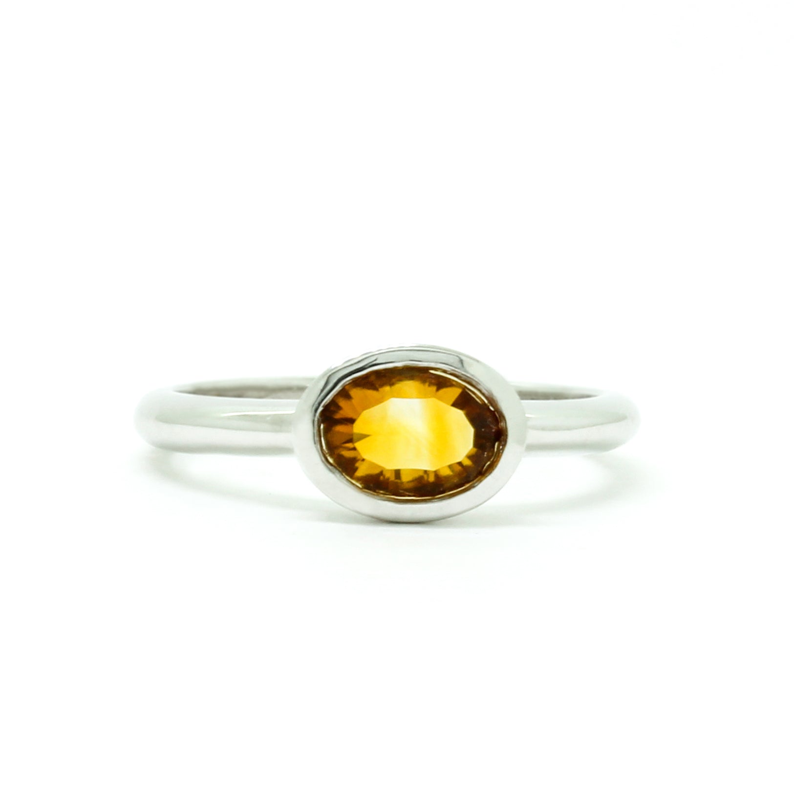 A product photo of a silver ring with a bezel-set citrine centre stone sitting on a white background. The silver band is simple and smooth, connecting on either side of a horizontally-oriented oval-cut citrine stone surrounded by a solid frame of silver. The citrine jewel is almost honey-coloured, reflecting a warm orangey yellow colour across its multi-faceted surface.