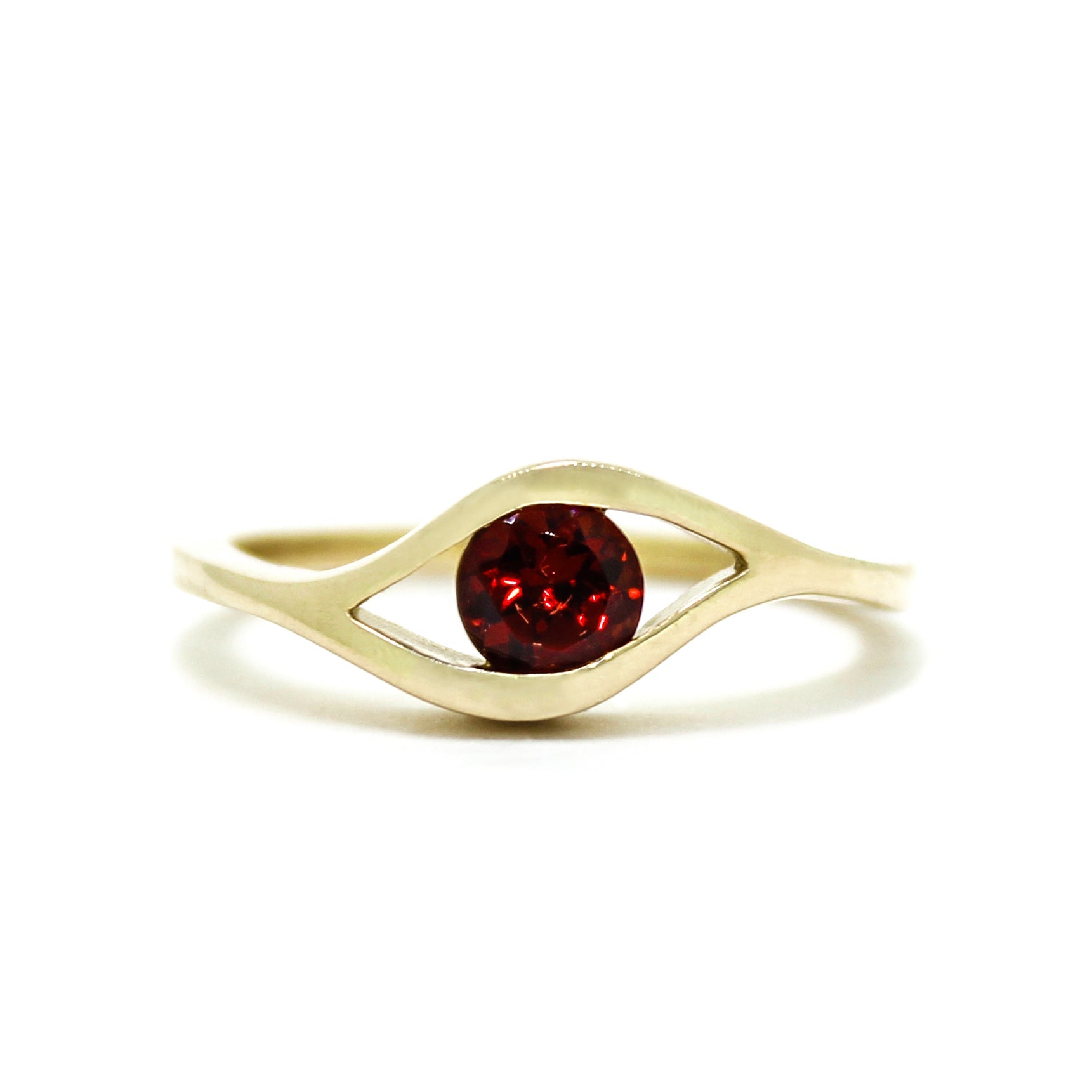 A product photo of a Round Garnet Ring in 9k Yellow Gold sitting on a plain white background. The 5.5mm garnet sits in the centre, and a simple golden band splits on either side of the jewel, meeting above and below, holding it in place like the corners of an eye. The stone is a deep red, reflecting sanguine hues across its multi-faceted edges.