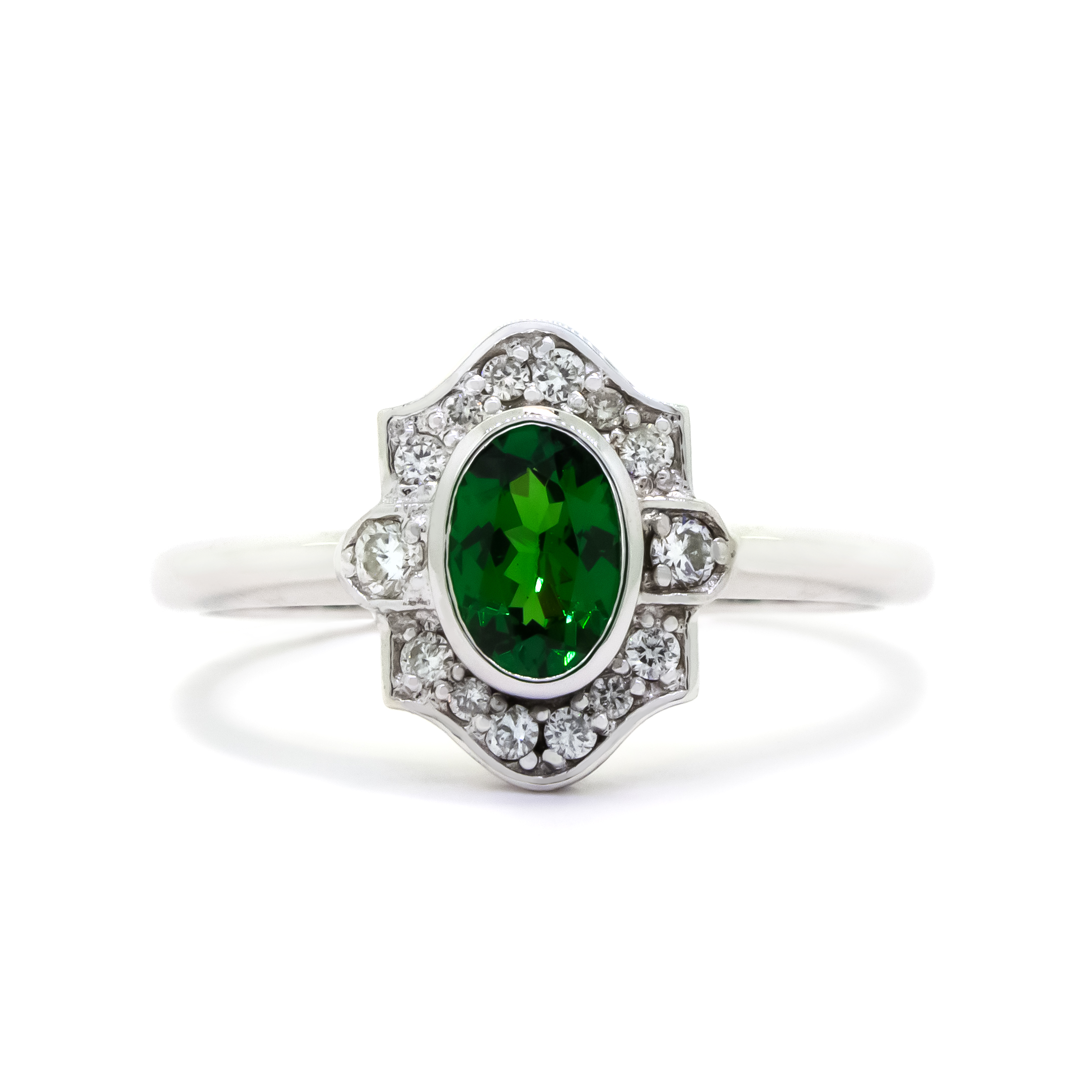 A product photo of a vintage-styled tsavorite and diamond ring in 9k white gold on a white background. The oval-shaped tsavorite is held in place by a bezel setting, and is surrounded by an ornate white gold frame embedded with diamonds, reminiscent of vintage jewellery styles of the past. The green gemstone colour be a good emerald substitute.