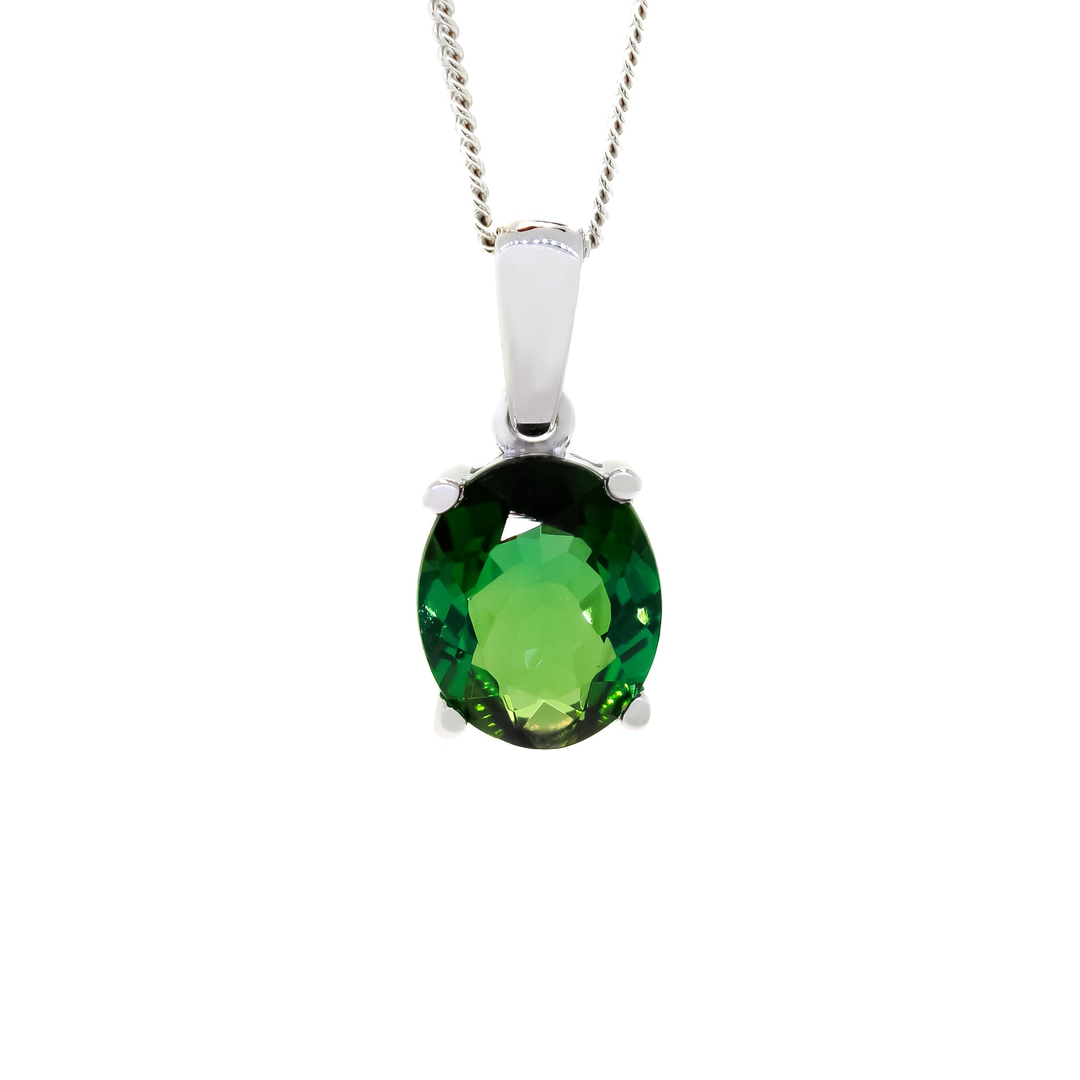 A product photo of a 9x7mm Oval Green Tourmaline Pendant in 9k White Gold suspended against a white background. The impressively large and deeply-coloured oval-cut stone is contrasted by its overall minimalistic design, 4 simple golden claws holding the gem in place. It is suspended by a simple gold chain. The stone is a deep green, reflecting verdant hues across its multi-faceted edges.