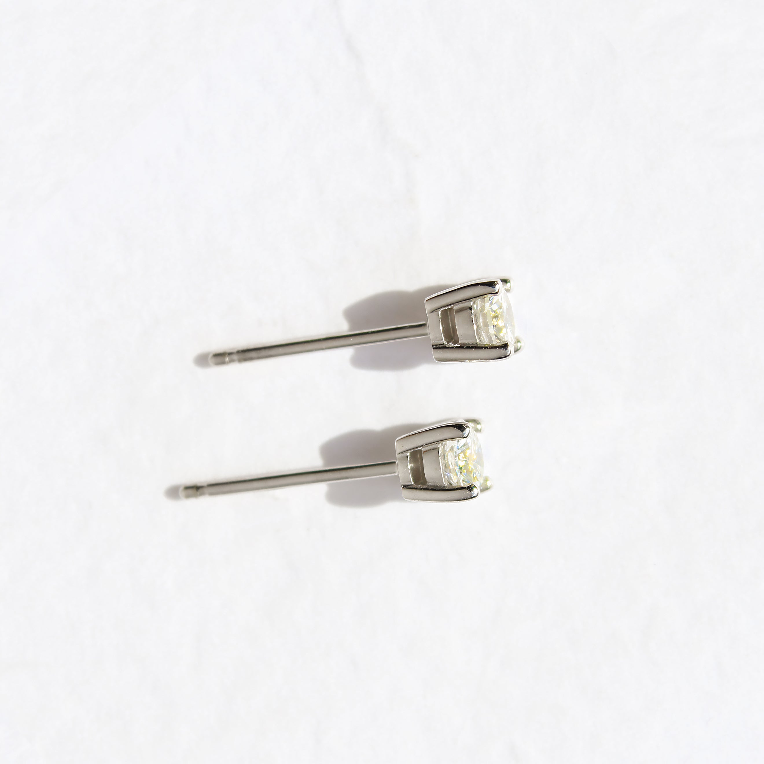 A product photo of a pair of 9 karat white gold lab diamond stud earrings sitting in the sun on a white textured background. The brilliant, colourless half pointer round diamonds measure approximately 4mm across, and are held in place by a simple 4-claw setting.