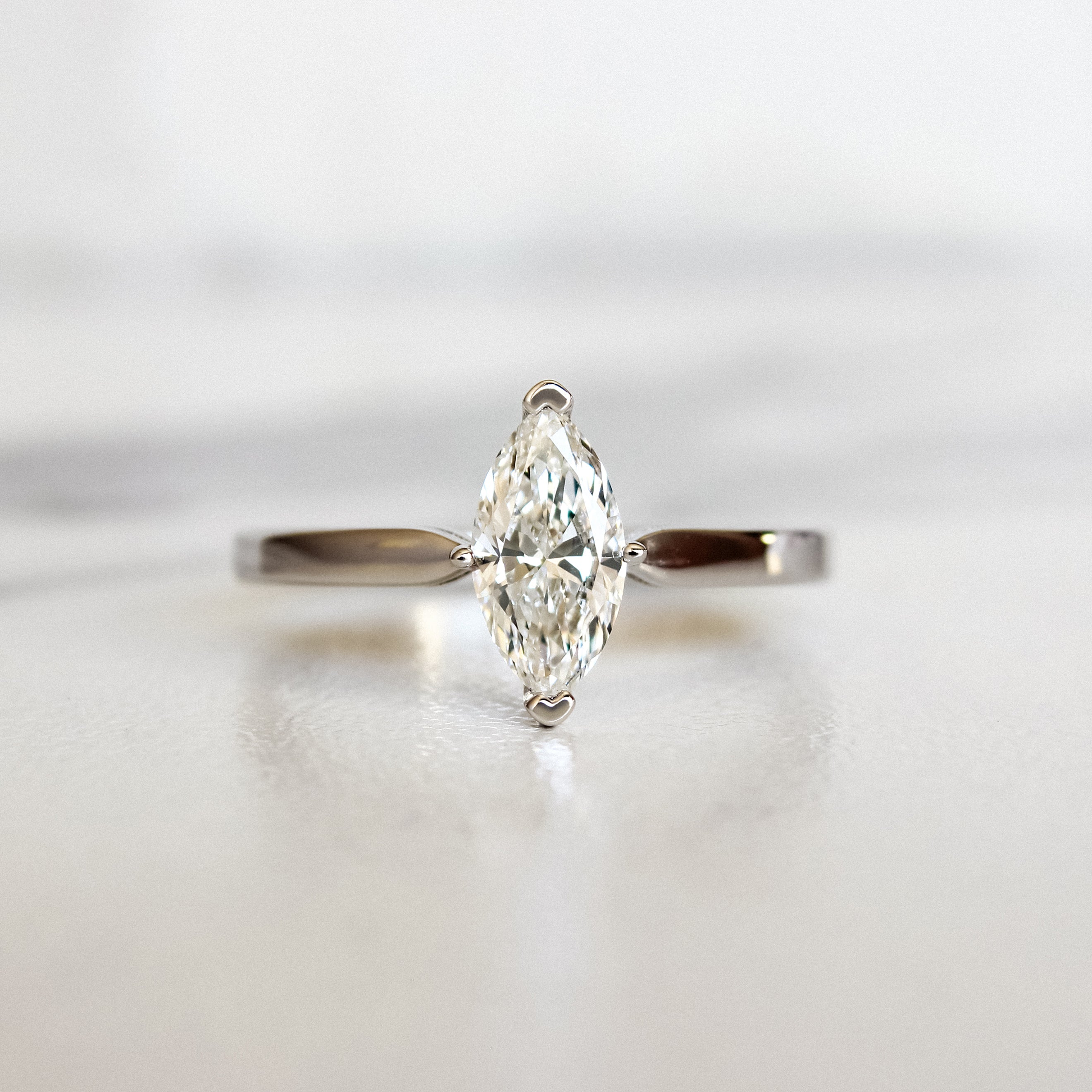 A product photo of a 14 karat white gold lab diamond solitaire engagement ring sitting in the sun on a white textured background. The brilliant, colourless 0.70ct marquise diamond measures about 8.5mm lengthwise, and is held in place by 4 delicate claws in a low-profile prong setting in the middle of a sharply tapered band.