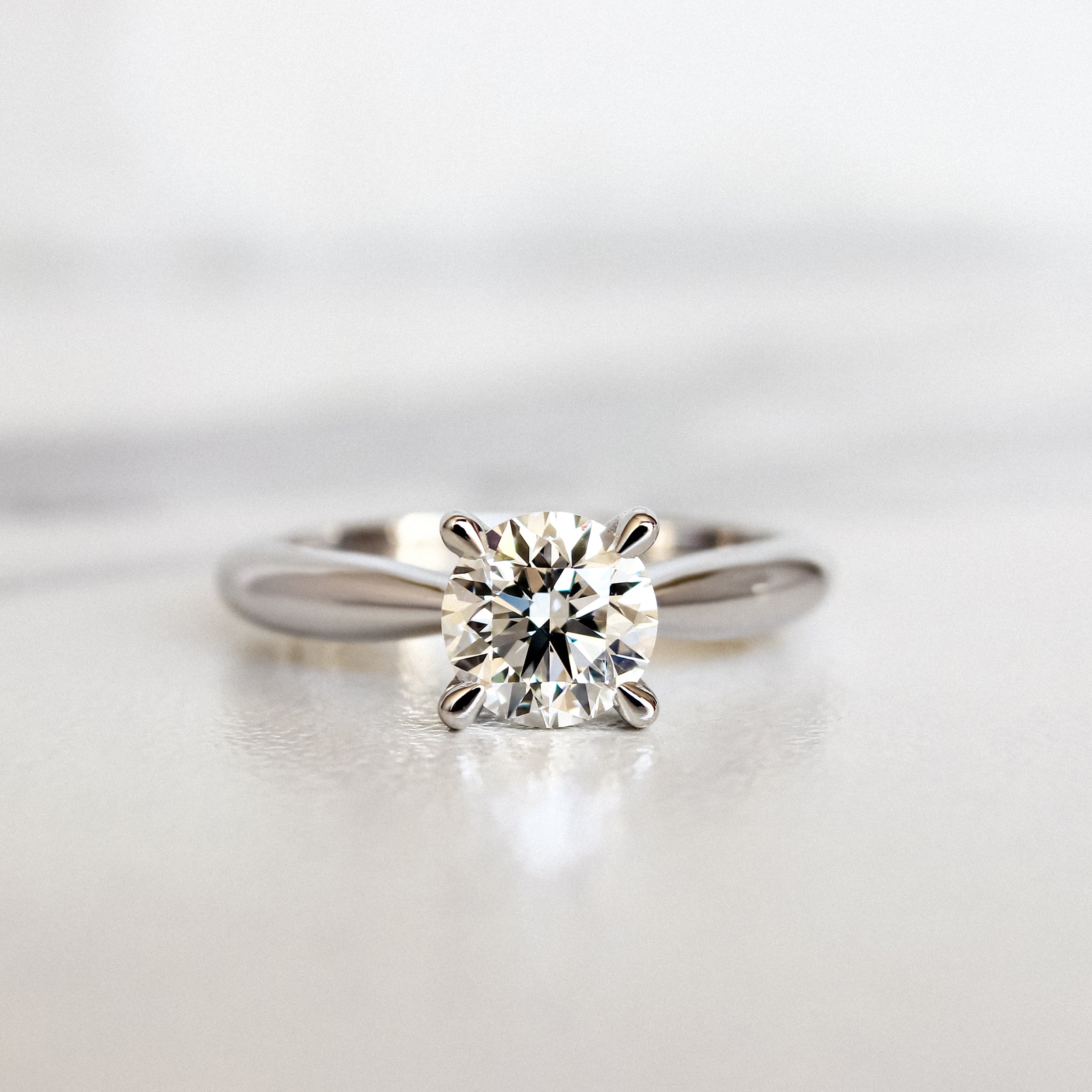 A product photo of a 14 karat white gold lab diamond solitaire engagement ring sitting in the sun on a white textured background. The brilliant 1 carat round-cut diamond measures 6.3mm lengthwise, and is held in place by 4 delicate claws in a prong setting in the middle of the thickest part of the gold band.