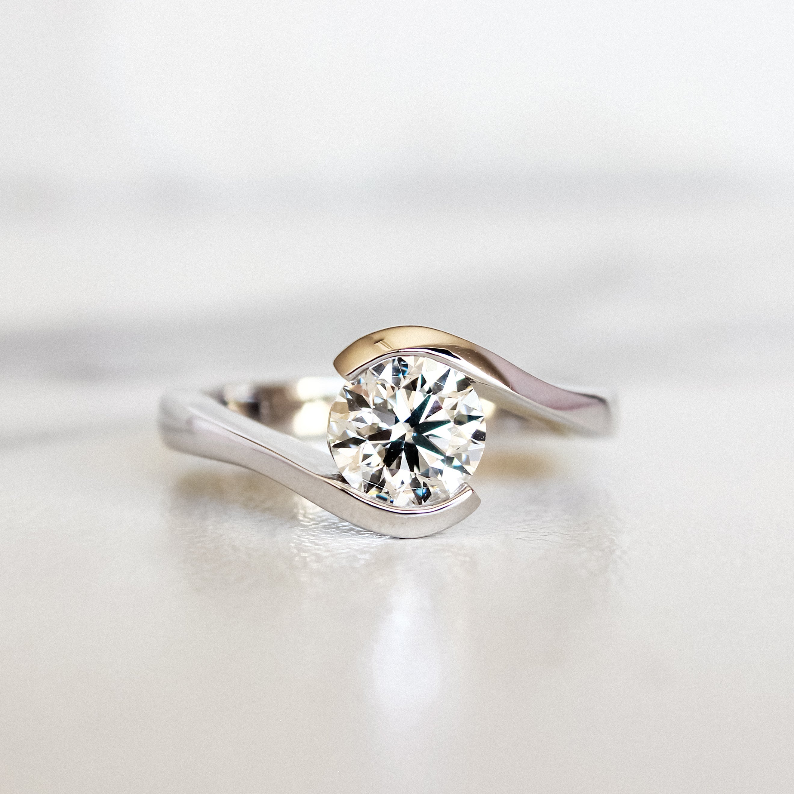 A product photo of a 14 karat white gold lab diamond solitaire engagement ring sitting in the sun on a white textured background. The brilliant, colourless 1 carat round-cut diamond measures 6.3mm lengthwise, and is held in place by a thick white gold band that curves around the top and bottom of the stone in a tension setting.