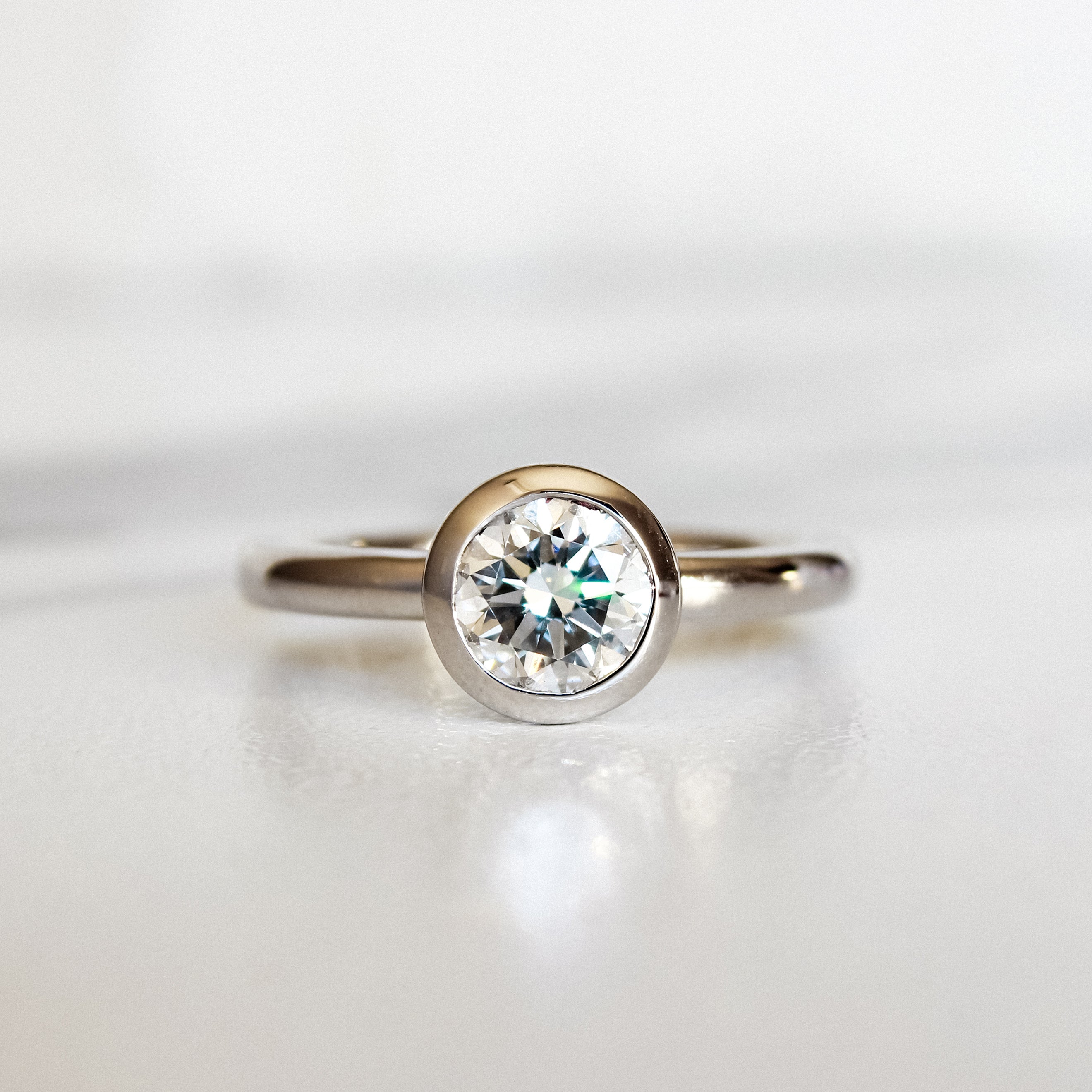 A product photo of a 14 karat white gold lab diamond solitaire engagement ring sitting in the sun on a white textured background. The brilliant, colourless 1 carat round-cut diamond measures 6.3mm lengthwise, and is held in place by a thick frame of white gold in a bezel setting.