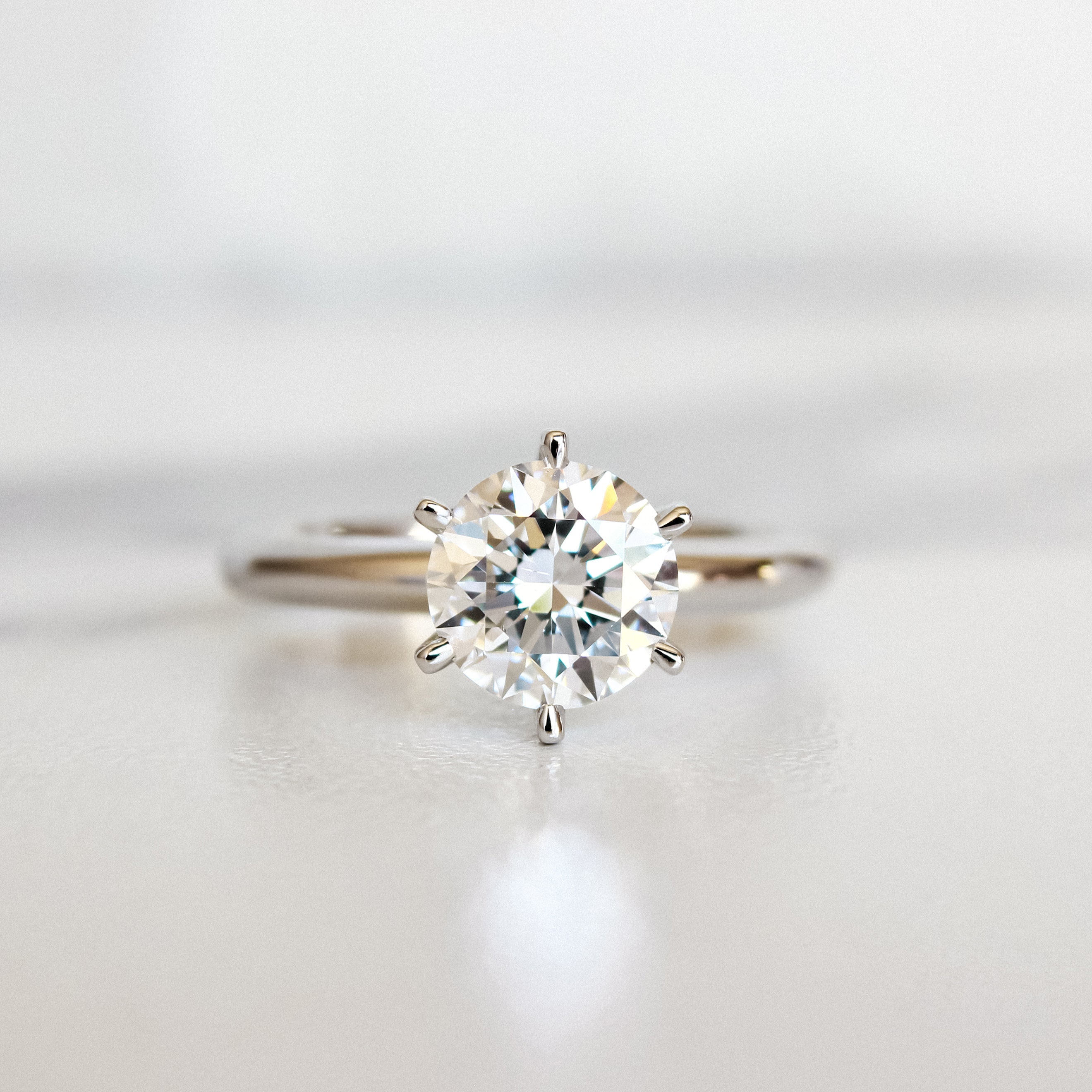 A product photo of a 14 karat white gold lab diamond solitaire engagement ring sitting in the sun on a white textured background. The brilliant, colourless 1.5 carat round-cut diamond measures 7.4mm lengthwise, and is held in place by 6 delicate claws in a high-profile prong setting.